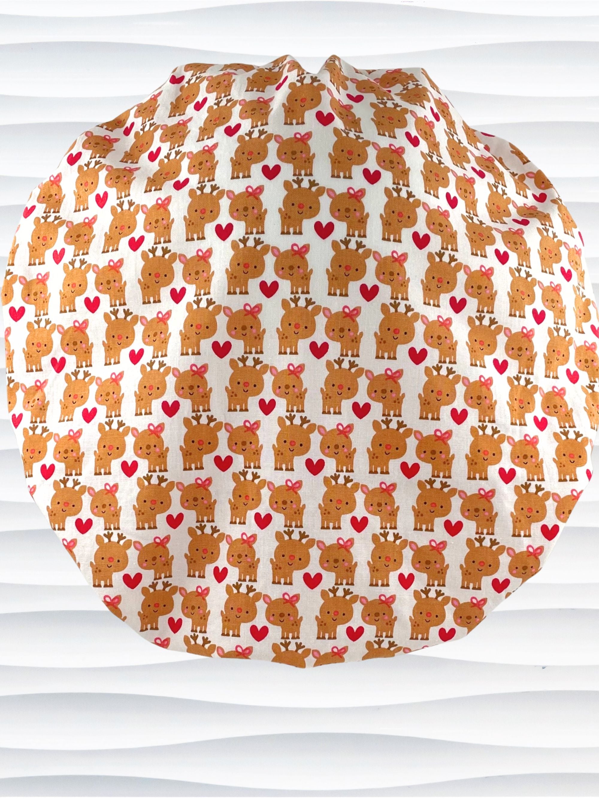 Bouffant style srugical scrub cap by Jenscaps made of white fabric patterned with repeating cute cartoon reindeer—some with pink bows—and red hearts, displayed against a white textured background.