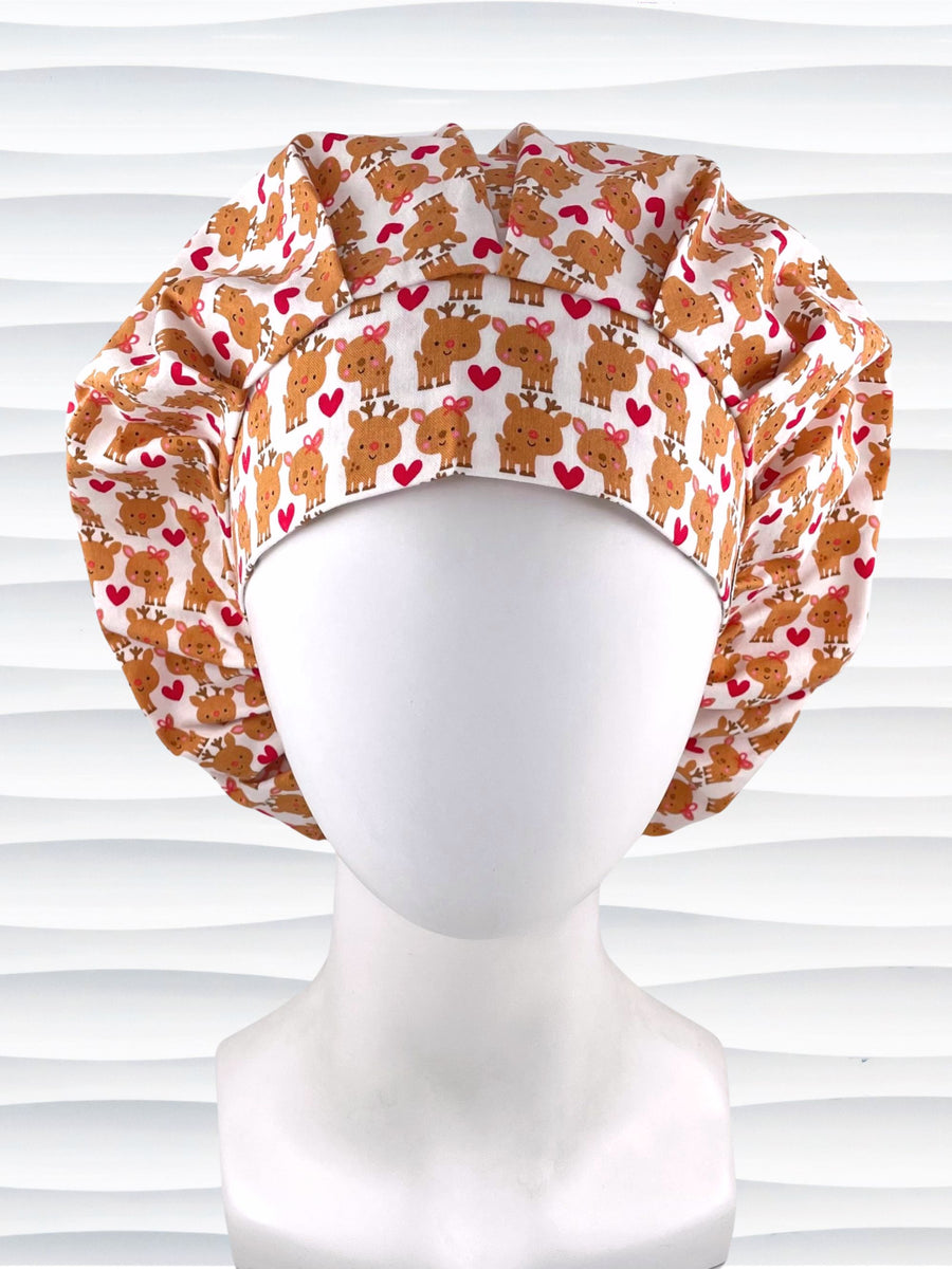 Bouffant style srugical scrub cap by Jenscaps made of white fabric patterned with repeating cute cartoon reindeer—some with pink bows—and red hearts, displayed against a white textured background.
