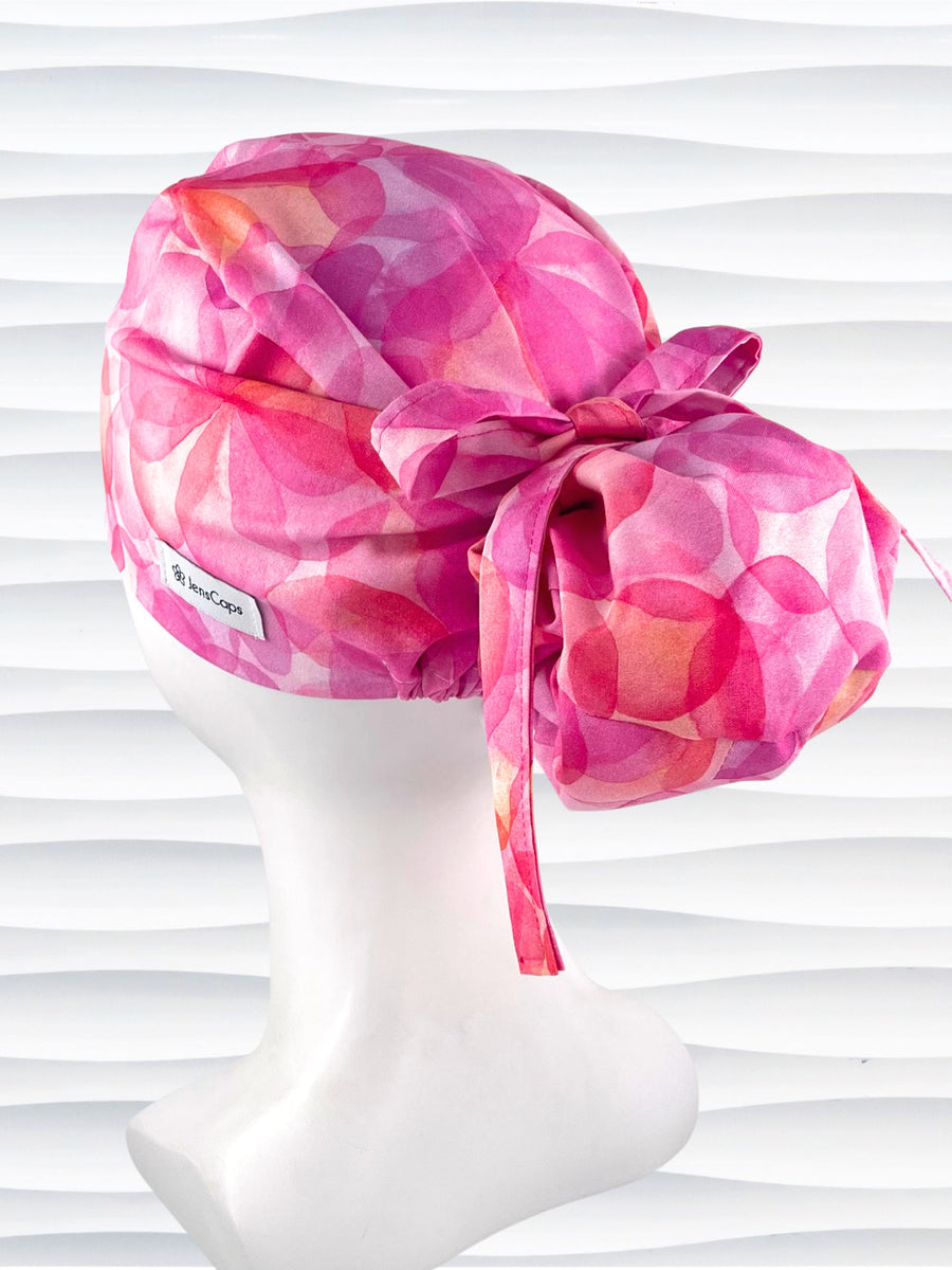 Euro Pony hybrid ponytail style surgical scrub cap by JensCaps with large flowers in light and dark pinks and peach sorbet on cotton fabric, finished with a ribbon tie and displayed on a mannequin head.