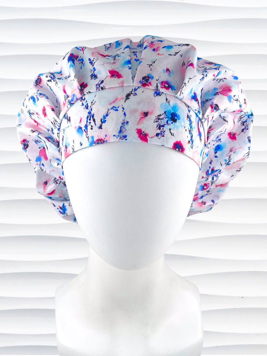 Bouffant style surgical scrub cap with watercolor looking pink and blue flowers and stems on white cotton fabric.