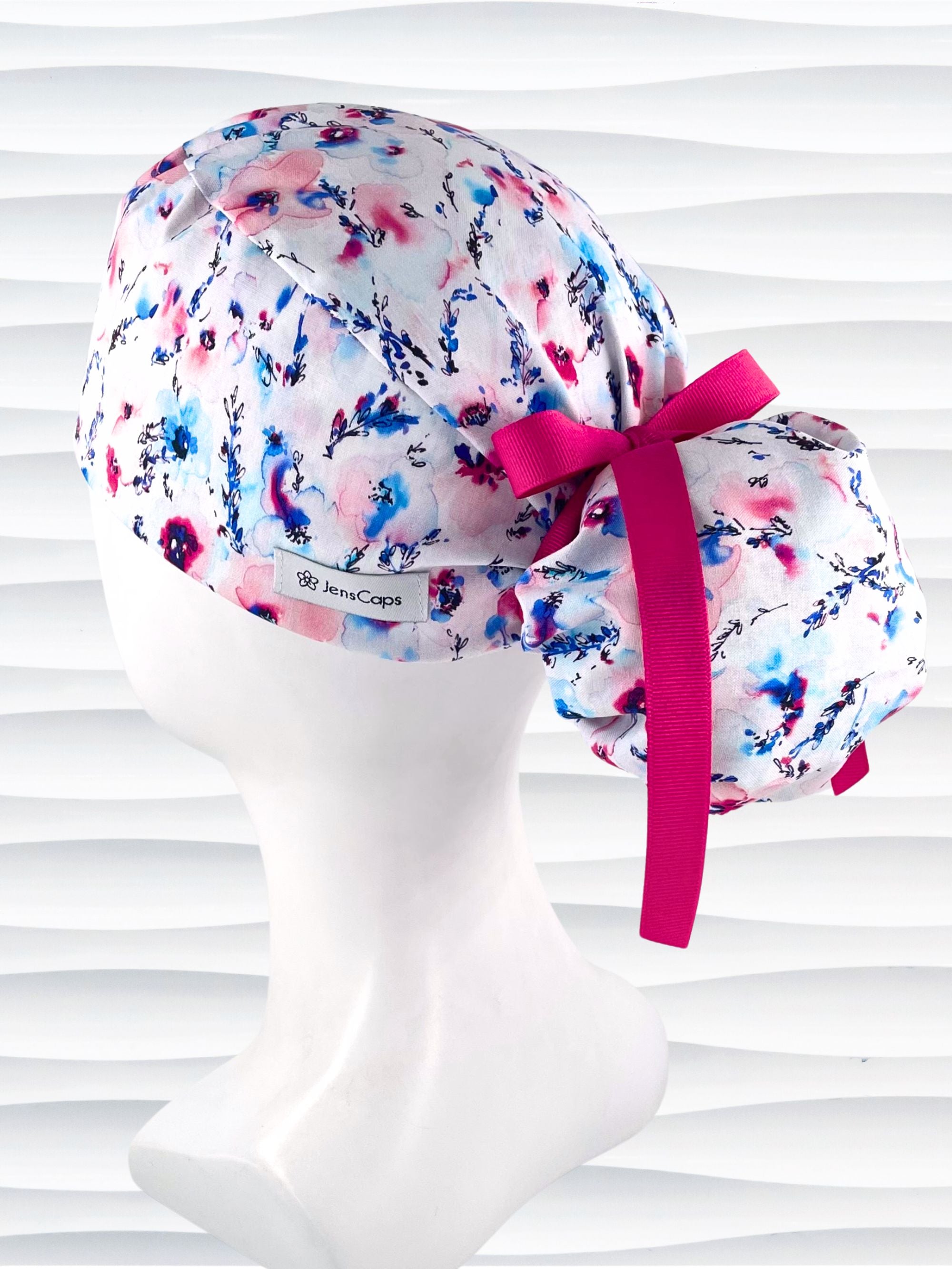 Ponytail style surgical scrub cap with abstract flowers in pinks and blues all over white premium cotton fabric and finished with grosgrain ribbon ties.
