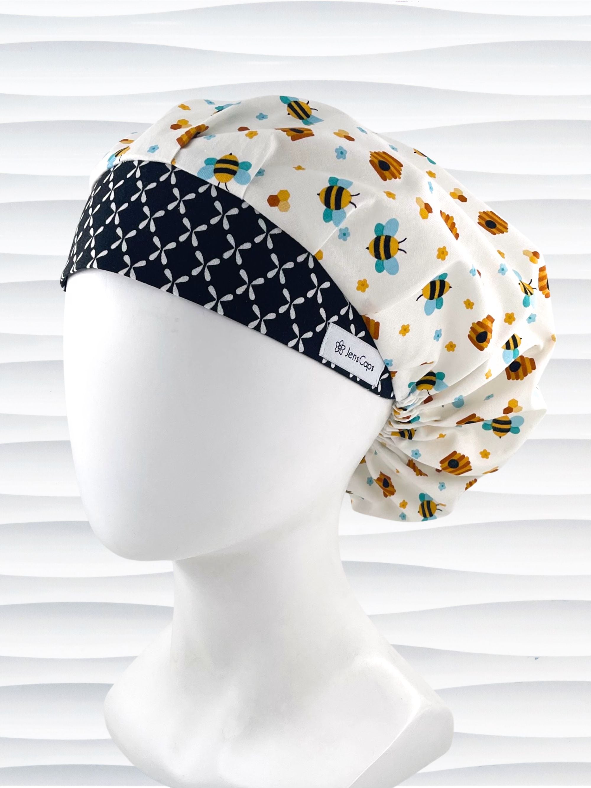 Bouffant style surgical scrub cap with an allover print of bumble bees, beehives, honeycombs, and flowers on white cotton fabric and finished with a black and white cooordinating print at the front.