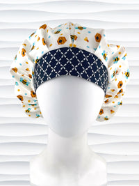 Bouffant style surgical scrub cap with an allover print of bumble bees, beehives, honeycombs, and flowers on white cotton fabric and finished with a black and white cooordinating print at the front.