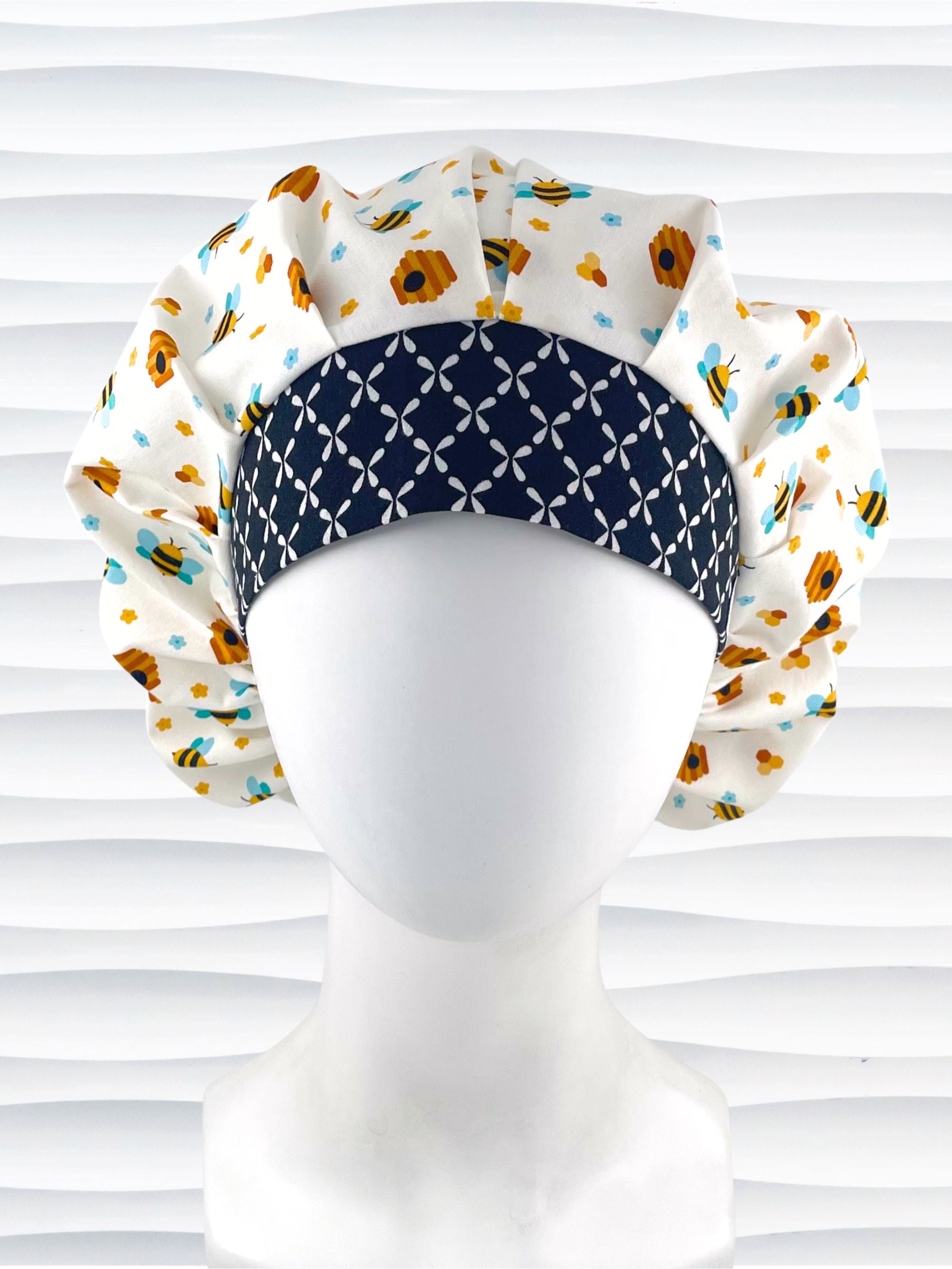 Bouffant style surgical scrub cap with an allover print of bumble bees, beehives, honeycombs, and flowers on white cotton fabric and finished with a black and white cooordinating print at the front.