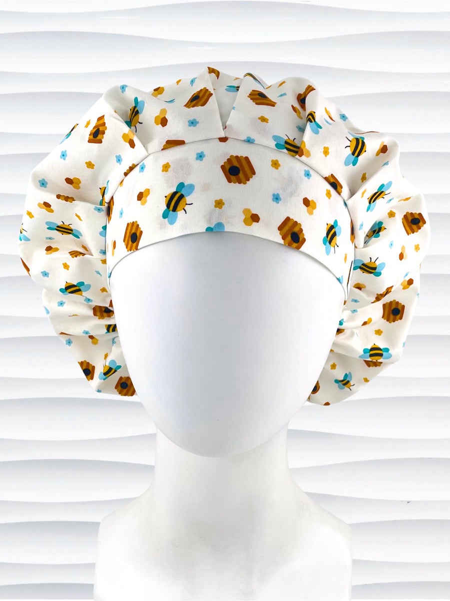 Bouffant style surgical scrub cap with an allover print of bumble bees, beehives, honeycombs, and flowers on white cotton fabric 