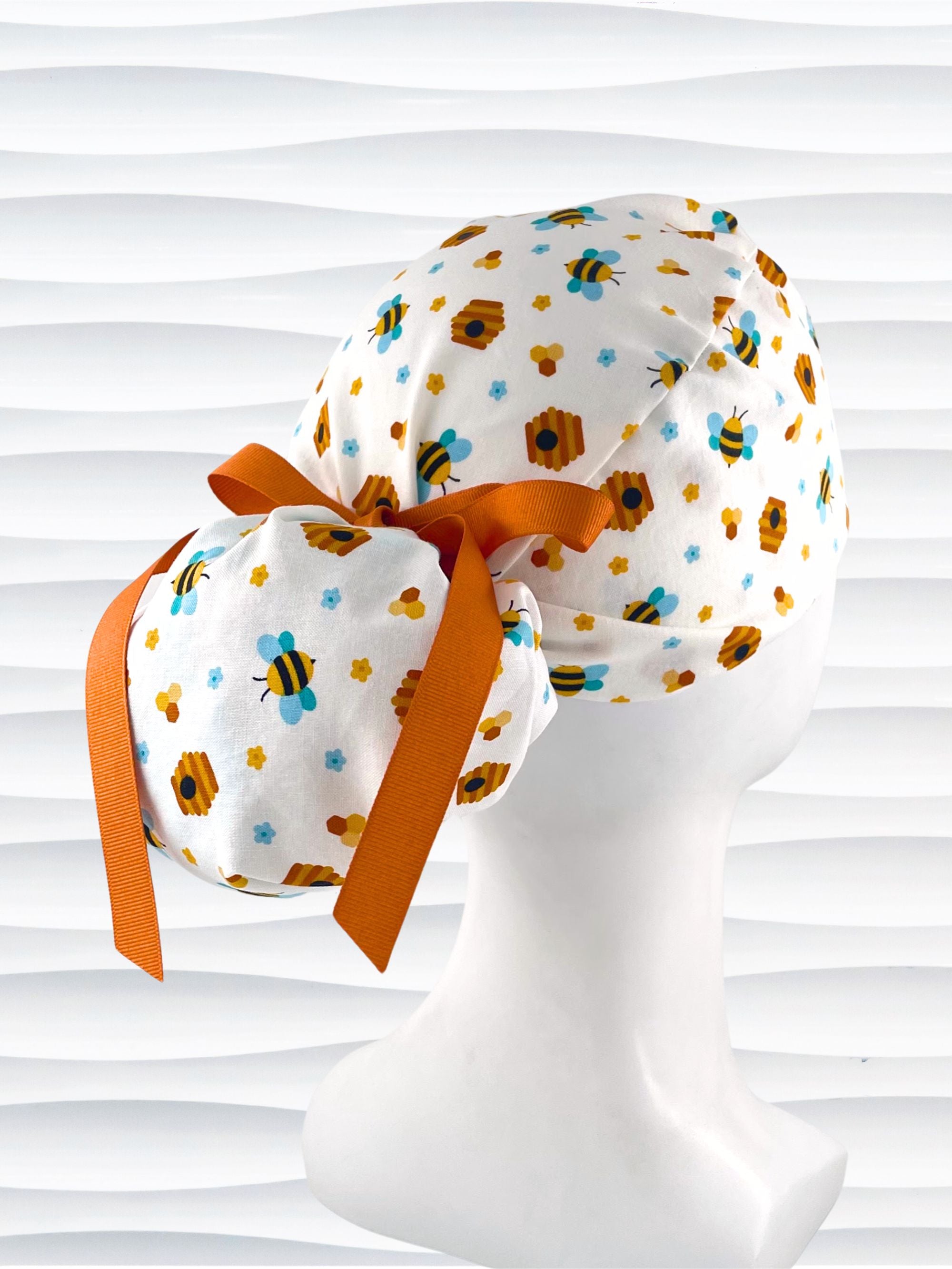 Ponytail style surgical scrub cap with honey bees with blue wings, blue and yellow daisies, and bee hives scattered all over this cotton fabric and tied with a grosgrain ribbon.