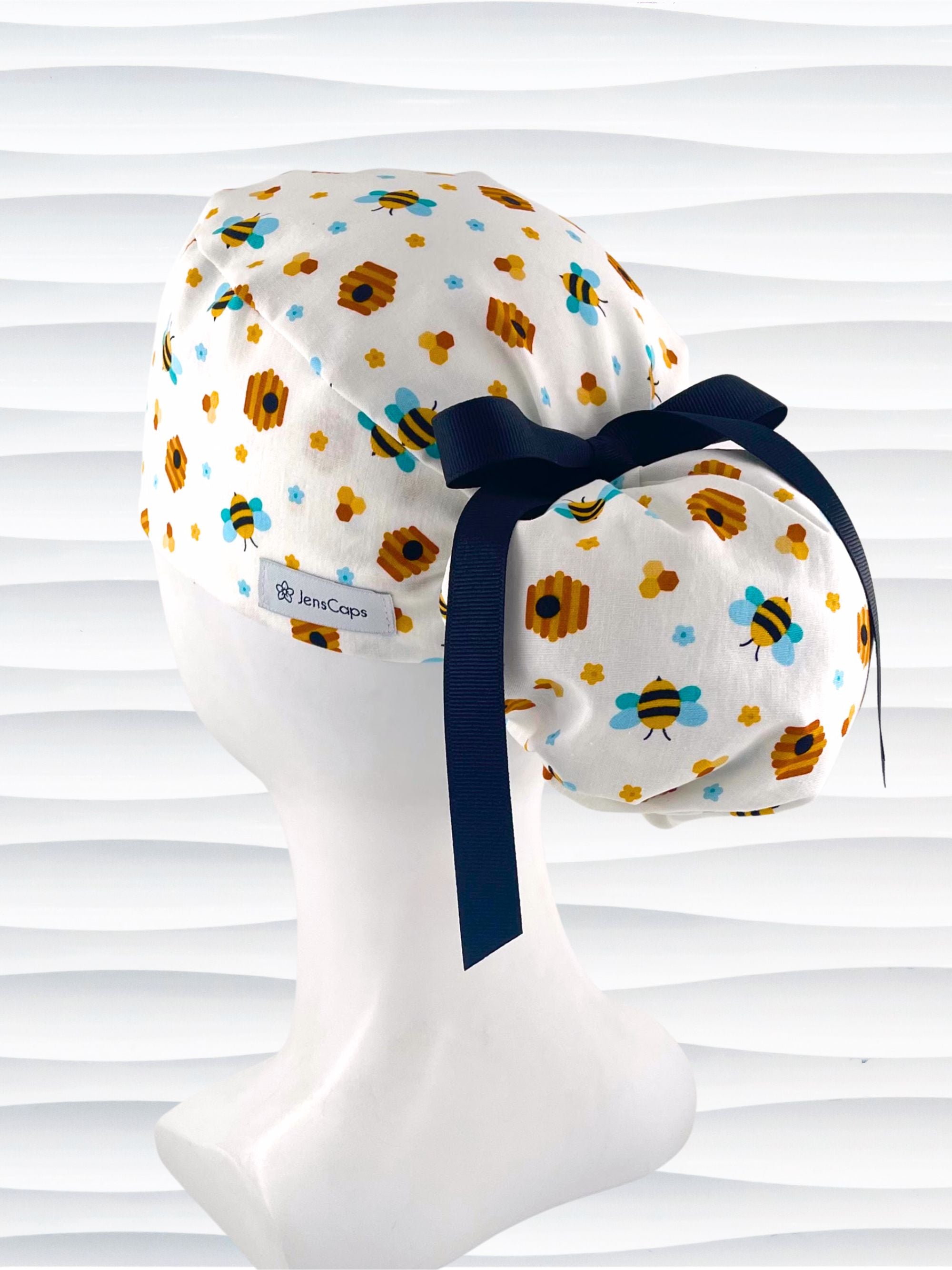 Ponytail style surgical scrub cap with honey bees with blue wings, blue and yellow daisies, and bee hives scattered all over this cotton fabric and tied with a grosgrain ribbon.