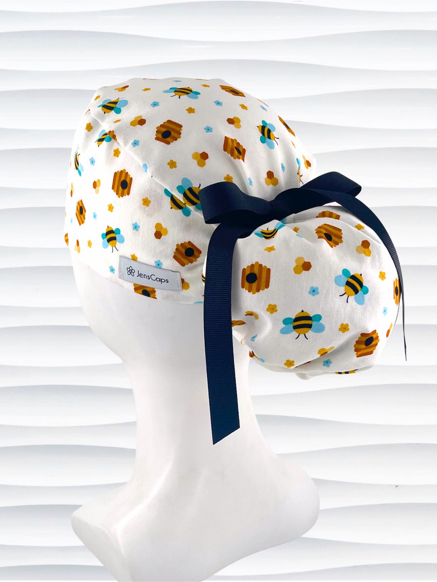 Ponytail style surgical scrub cap with honey bees with blue wings, blue and yellow daisies, and bee hives scattered all over this cotton fabric and tied with a grosgrain ribbon.