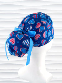 Pixie style surgical scrub cap with an allover print of country style red white and blue hearts with dots, stars, and stripes on royal blue cotton fabric.