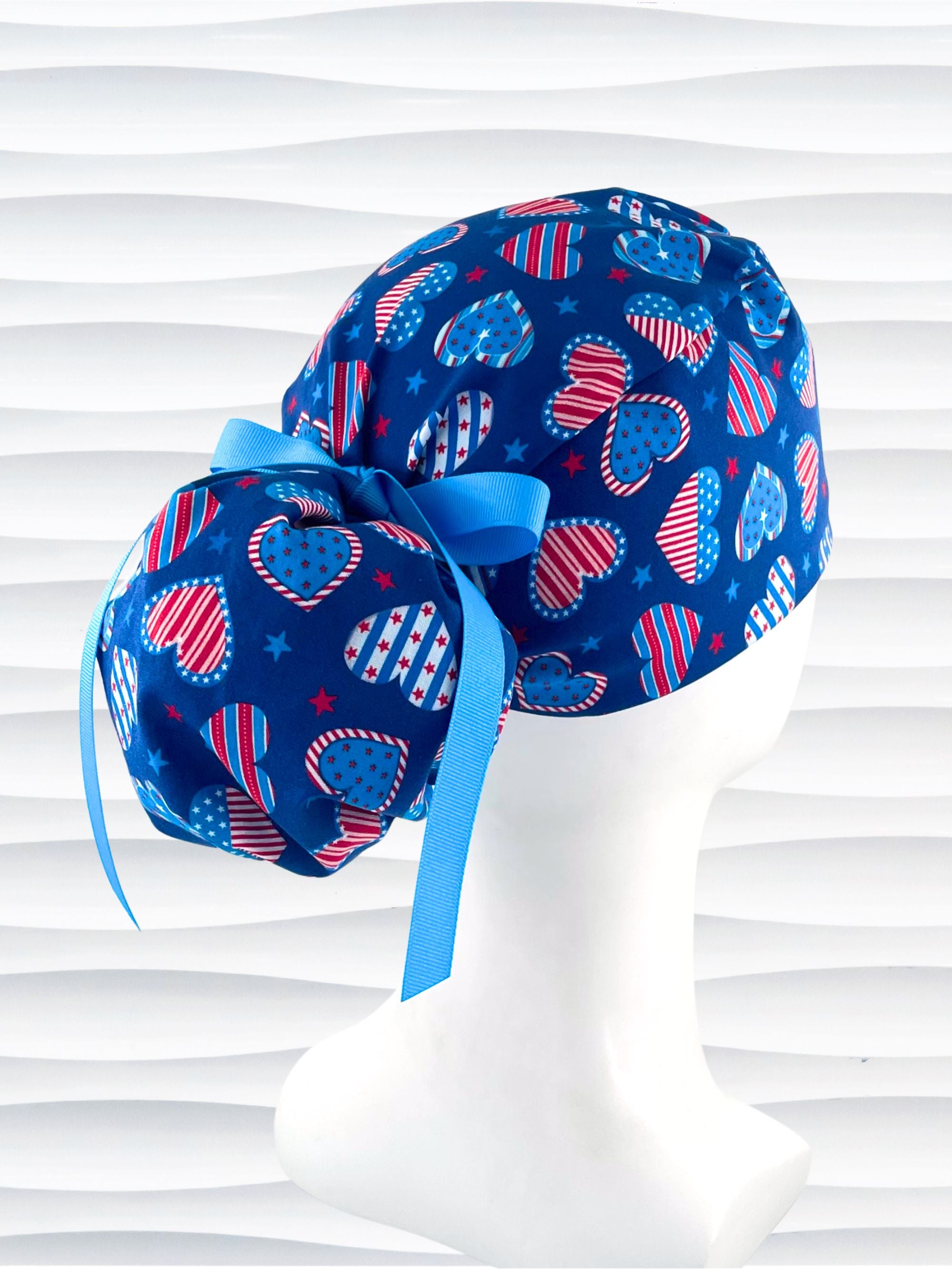 Pixie style surgical scrub cap with an allover print of country style red white and blue hearts with dots, stars, and stripes on royal blue cotton fabric.