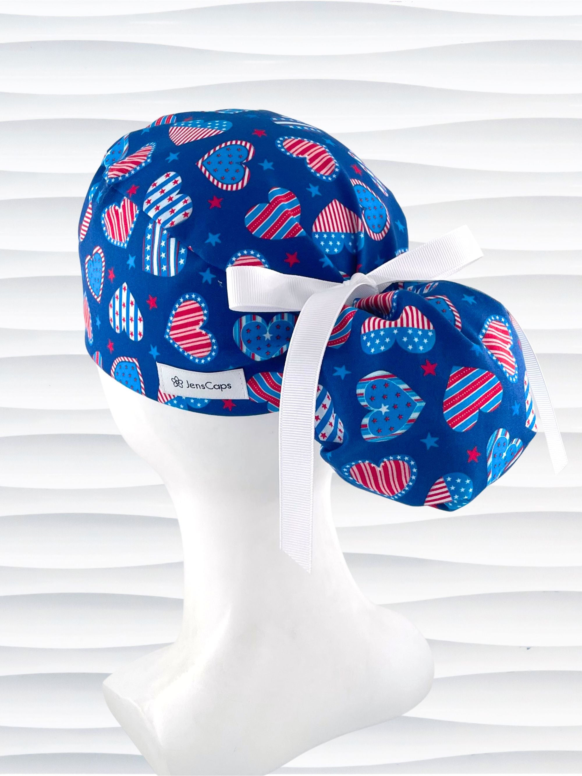 Pixie style surgical scrub cap with an allover print of country style red white and blue hearts with dots, stars, and stripes on royal blue cotton fabric.