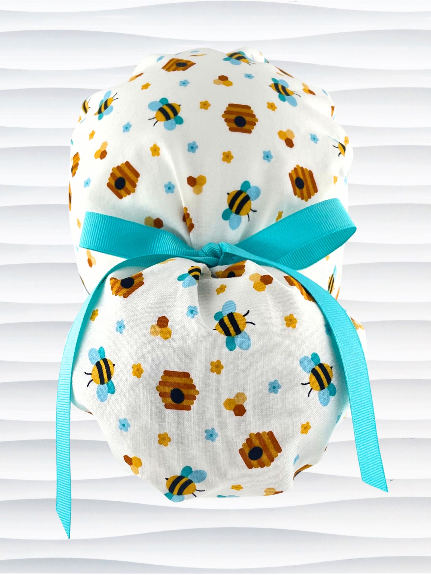 Ponytail style surgical scrub cap with honey bees with blue wings, blue and yellow daisies, and bee hives scattered all over this cotton fabric and tied with a grosgrain ribbon.