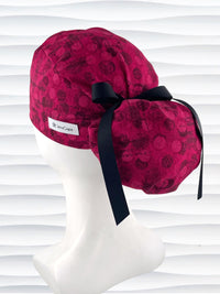 Ponytail pouch style surgical scrub cap with a muted pattern of black circles and lines on deep red or magenta cotton fabric.