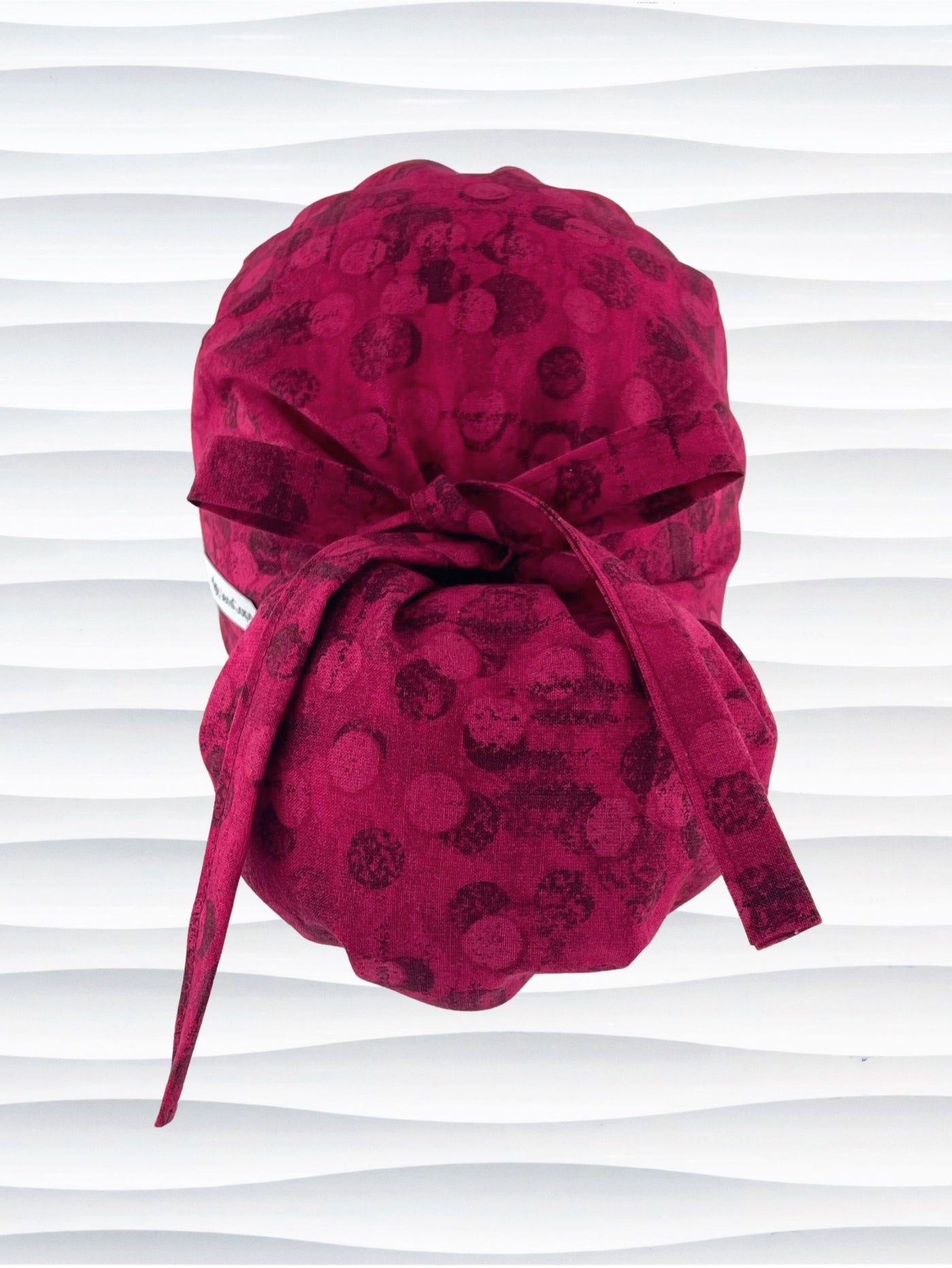 Ponytail pouch style surgical scrub cap with a muted pattern of black circles and lines on deep red or magenta cotton fabric.