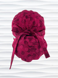Ponytail pouch style surgical scrub cap with a muted pattern of black circles and lines on deep red or magenta cotton fabric.