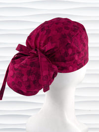Ponytail pouch style surgical scrub cap with a muted pattern of black circles and lines on deep red or magenta cotton fabric.