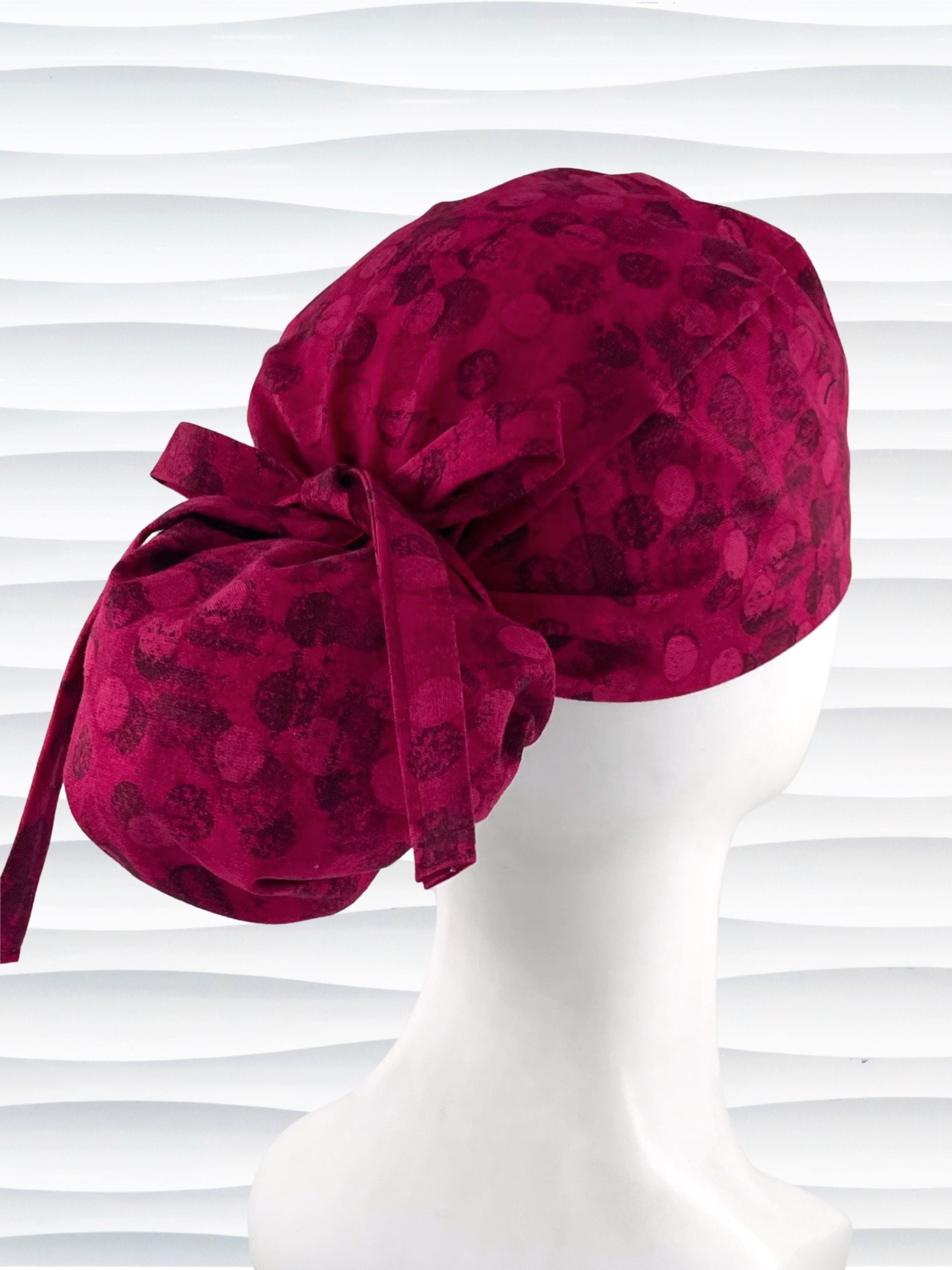 Ponytail pouch style surgical scrub cap with a muted pattern of black circles and lines on deep red or magenta cotton fabric.