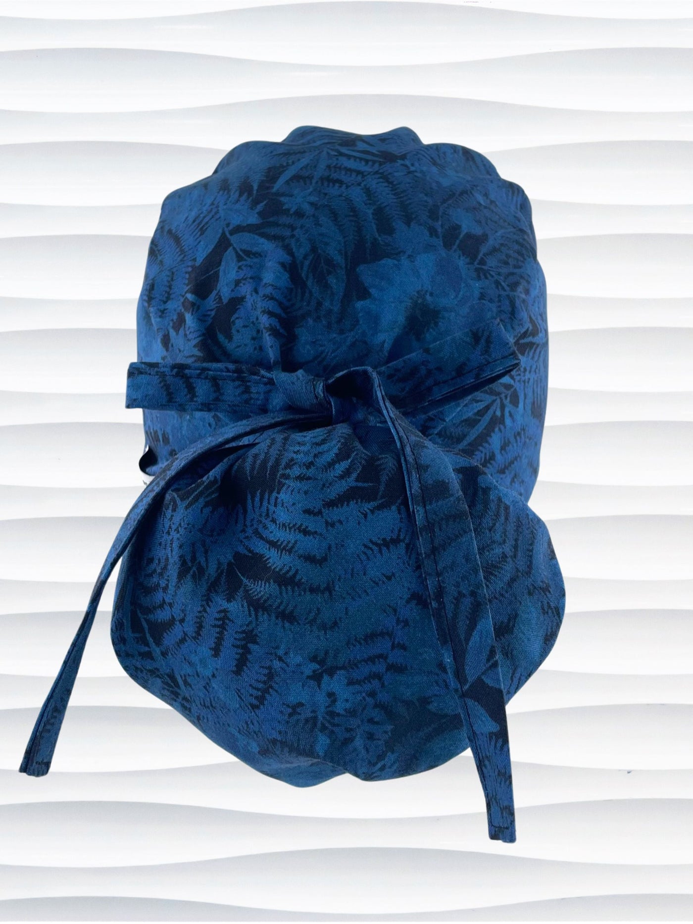 Ponytail pouch style surgical scrub cap with a muted pattern of flowers, ferns, and leaves in dark blue on a deep royal blue cotton fabric.