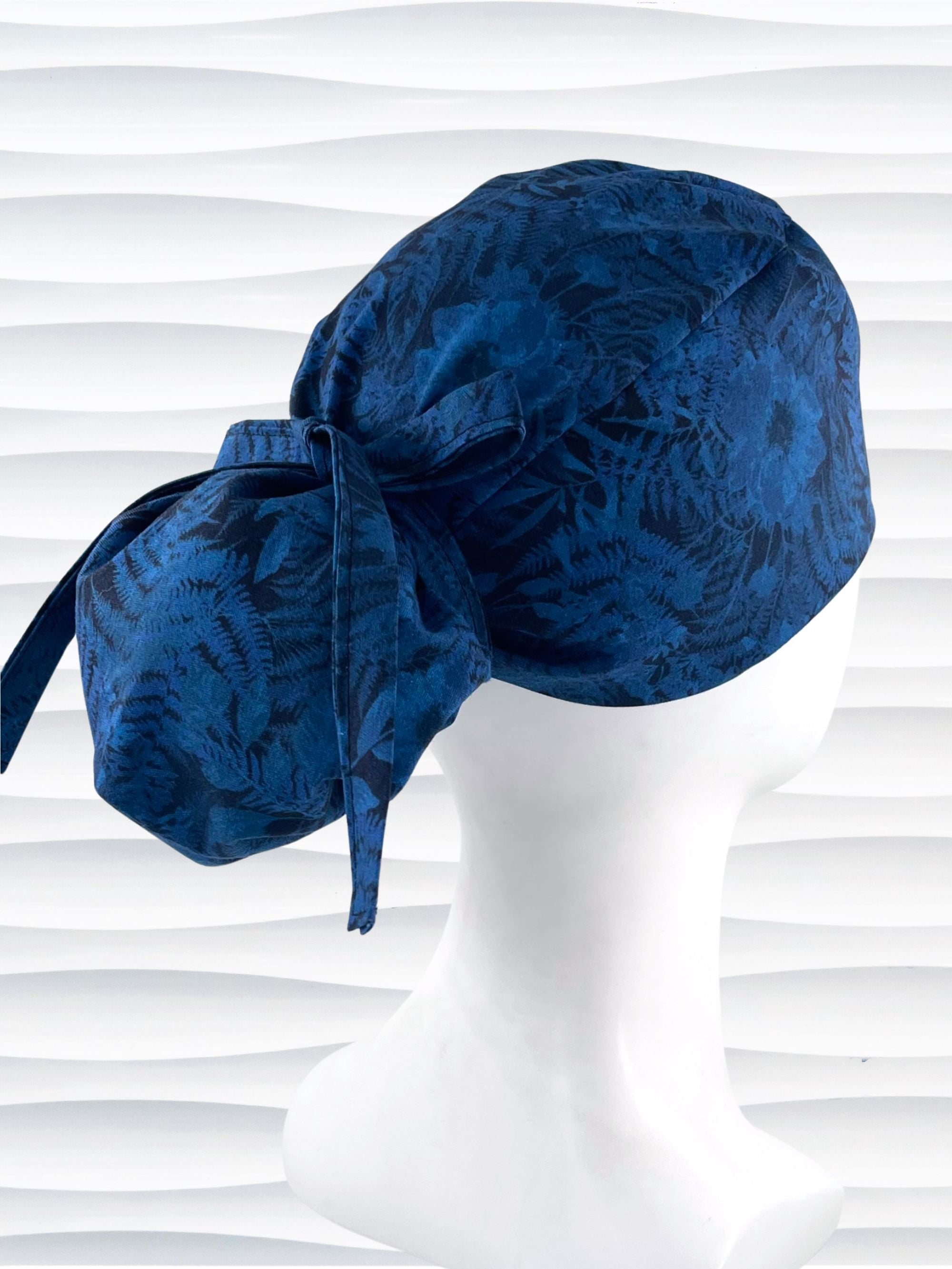 Ponytail pouch style surgical scrub cap with a muted pattern of flowers, ferns, and leaves in dark blue on a deep royal blue cotton fabric.
