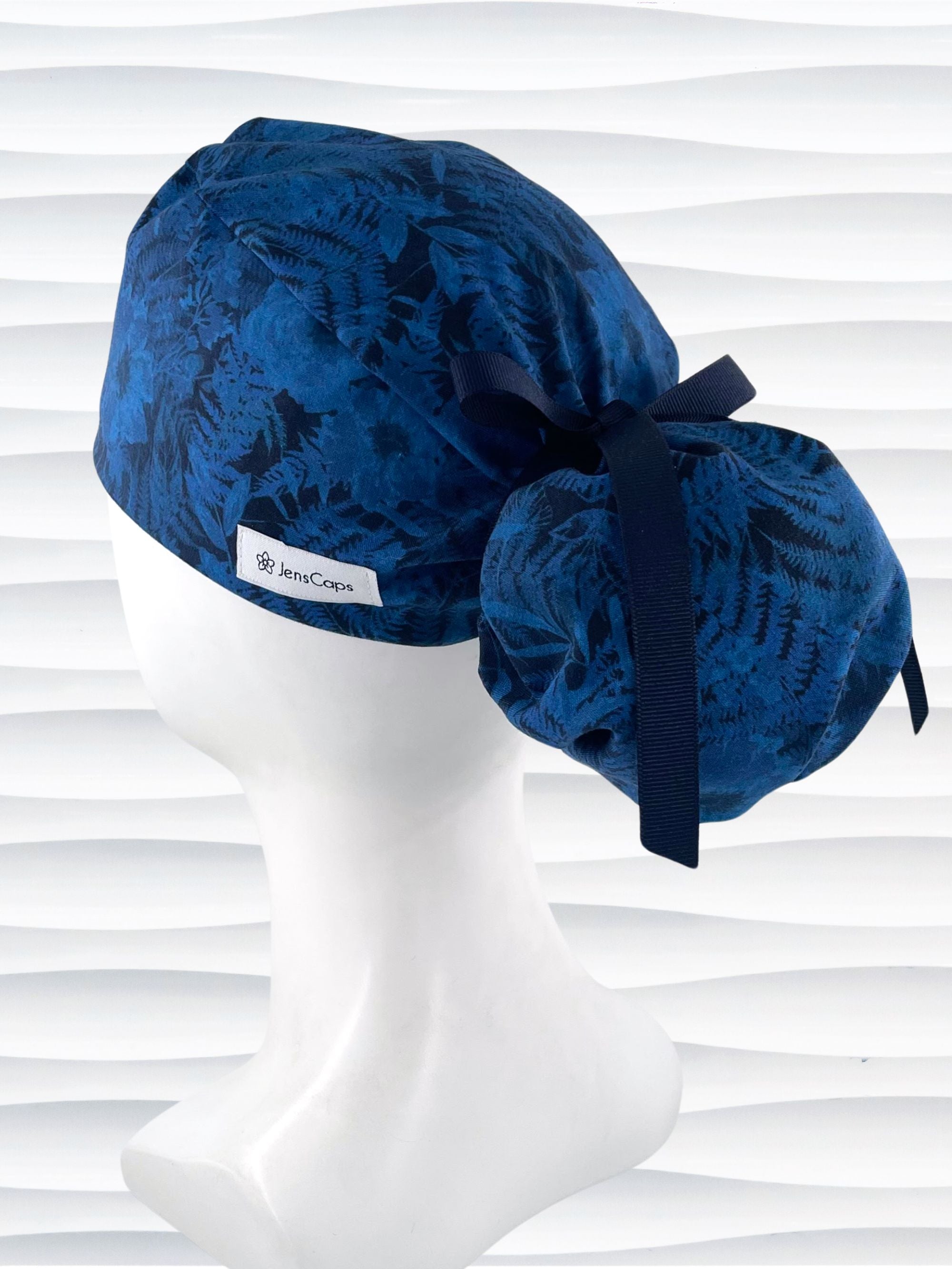 Ponytail pouch style surgical scrub cap with a muted pattern of flowers, ferns, and leaves in dark blue on a deep royal blue cotton fabric.