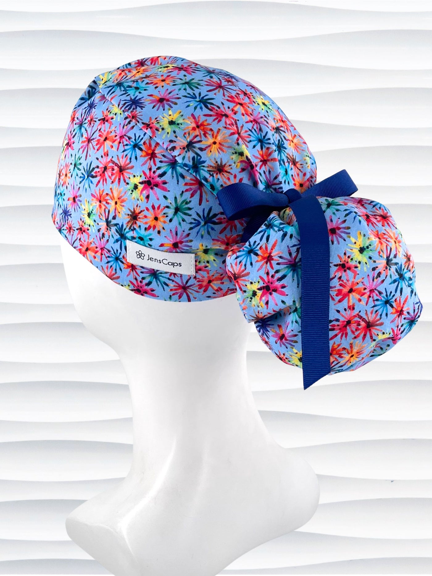 Ponytail pouch style surgical scrub cap with floral fireworks in pinks, reds, blues, yellow, and purple on cotton fabric and finished with a grosgrain ribbon tie.