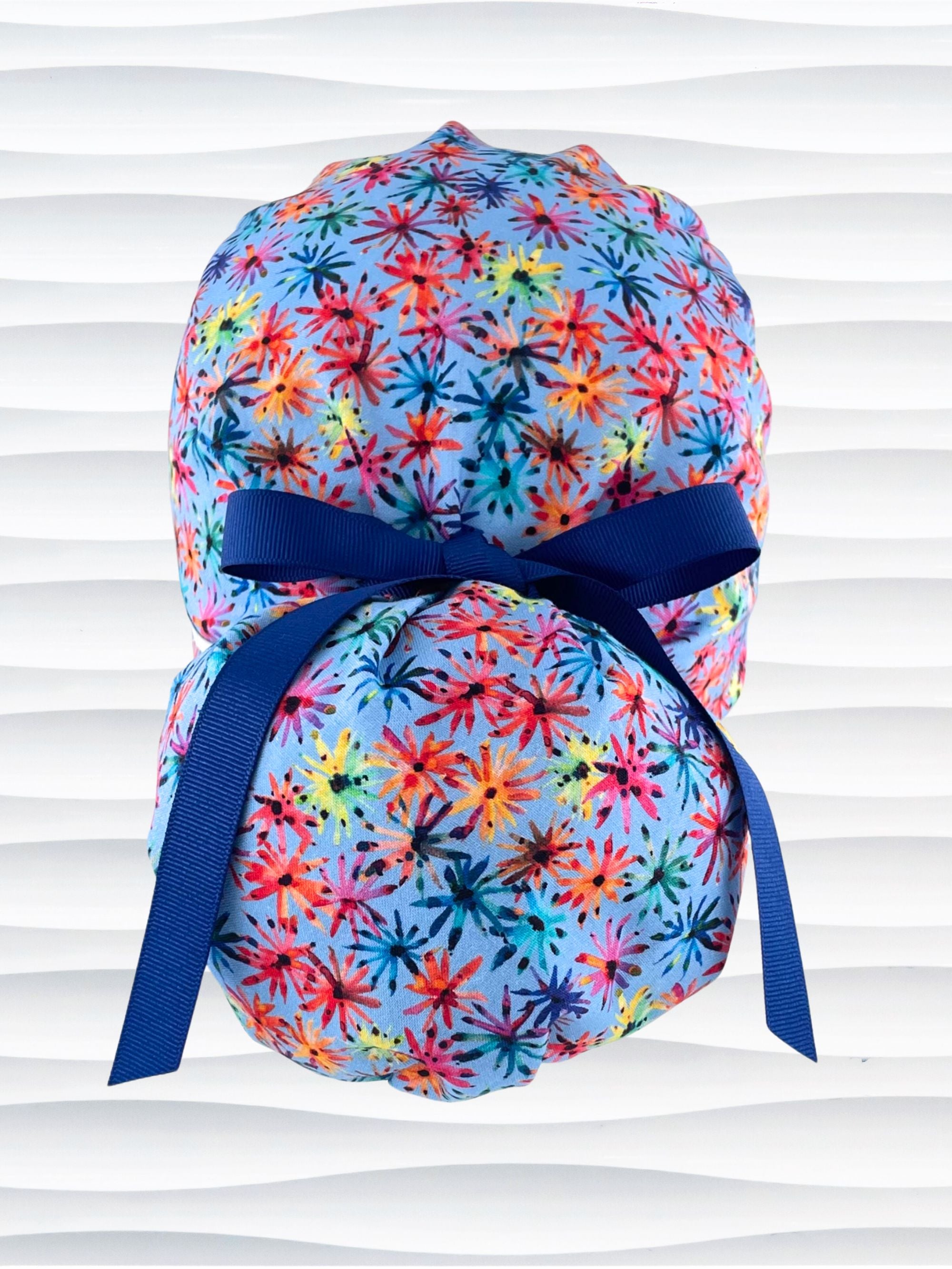 Ponytail pouch style surgical scrub cap with floral fireworks in pinks, reds, blues, yellow, and purple on cotton fabric and finished with a grosgrain ribbon tie.