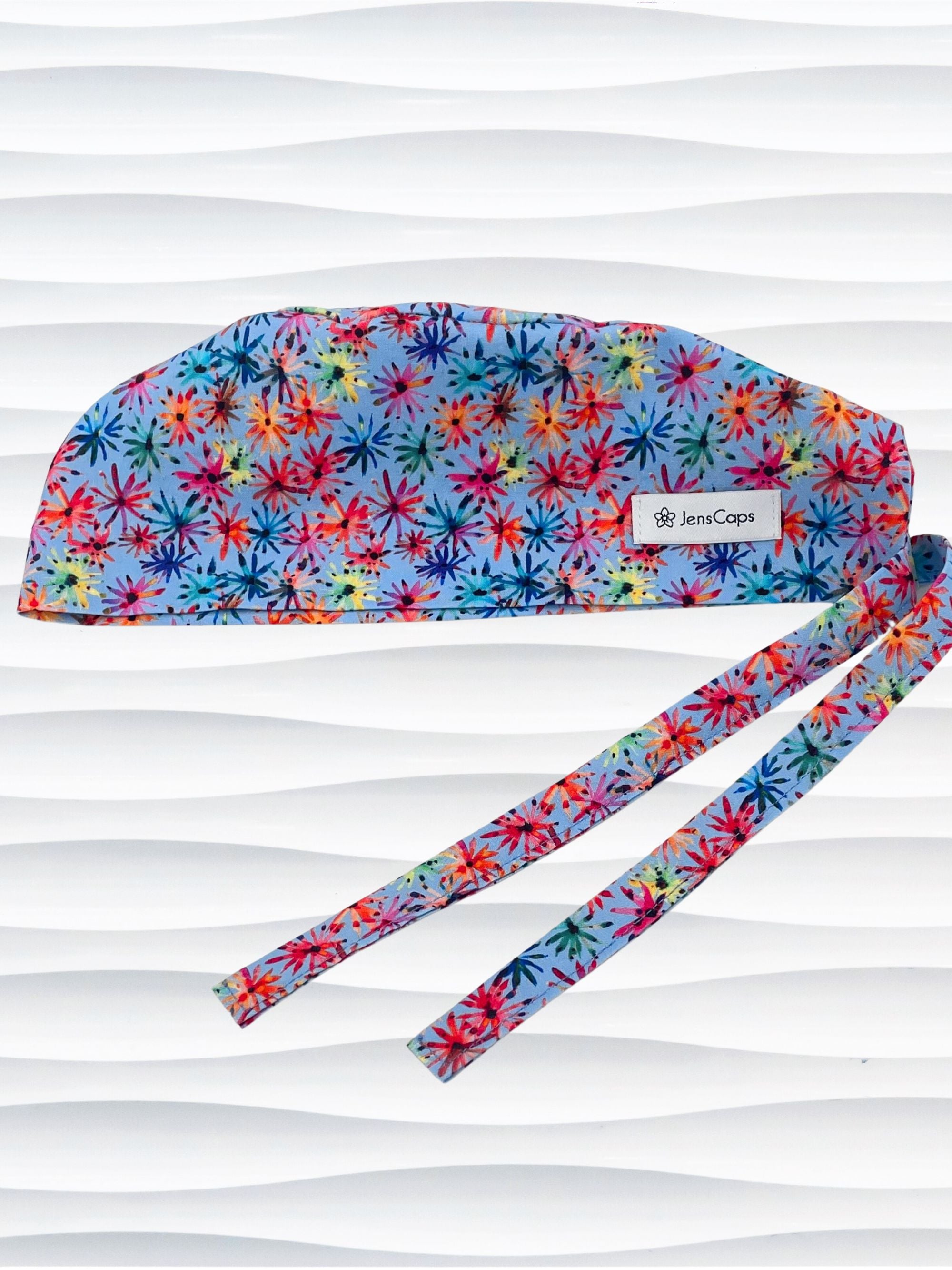 Surgeon style surgical scrub cap by JensCaps with red, blue, yellow, and green fireworks that look like flowers all over a medium blue background on cotton fabric.