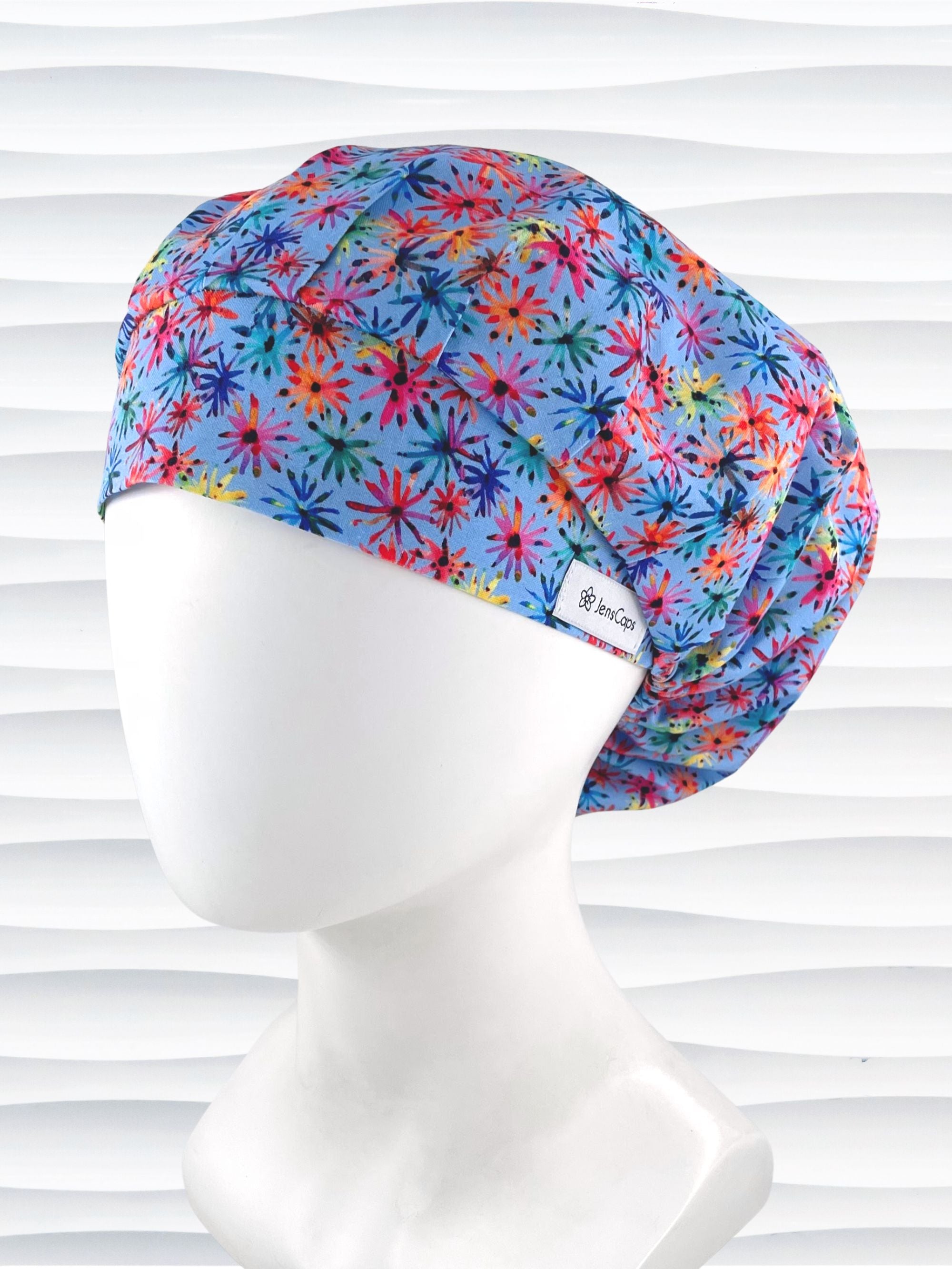Bouffant style surgical scrub cap with bright flowers that look like fireworks in red, pink, yellow, blue, and green all over blue cotton fabric.