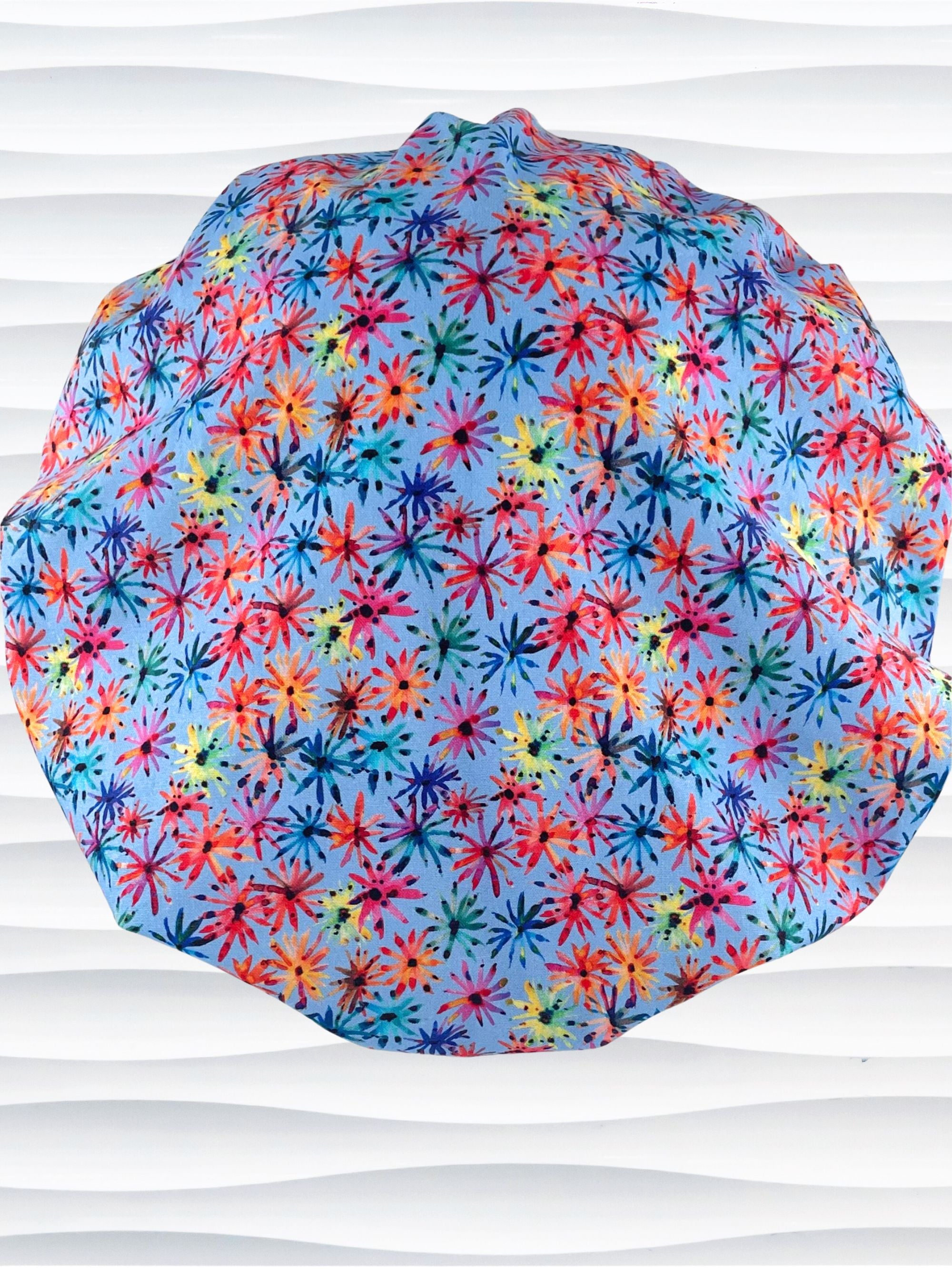 Bouffant style surgical scrub cap with bright flowers that look like fireworks in red, pink, yellow, blue, and green all over blue cotton fabric.