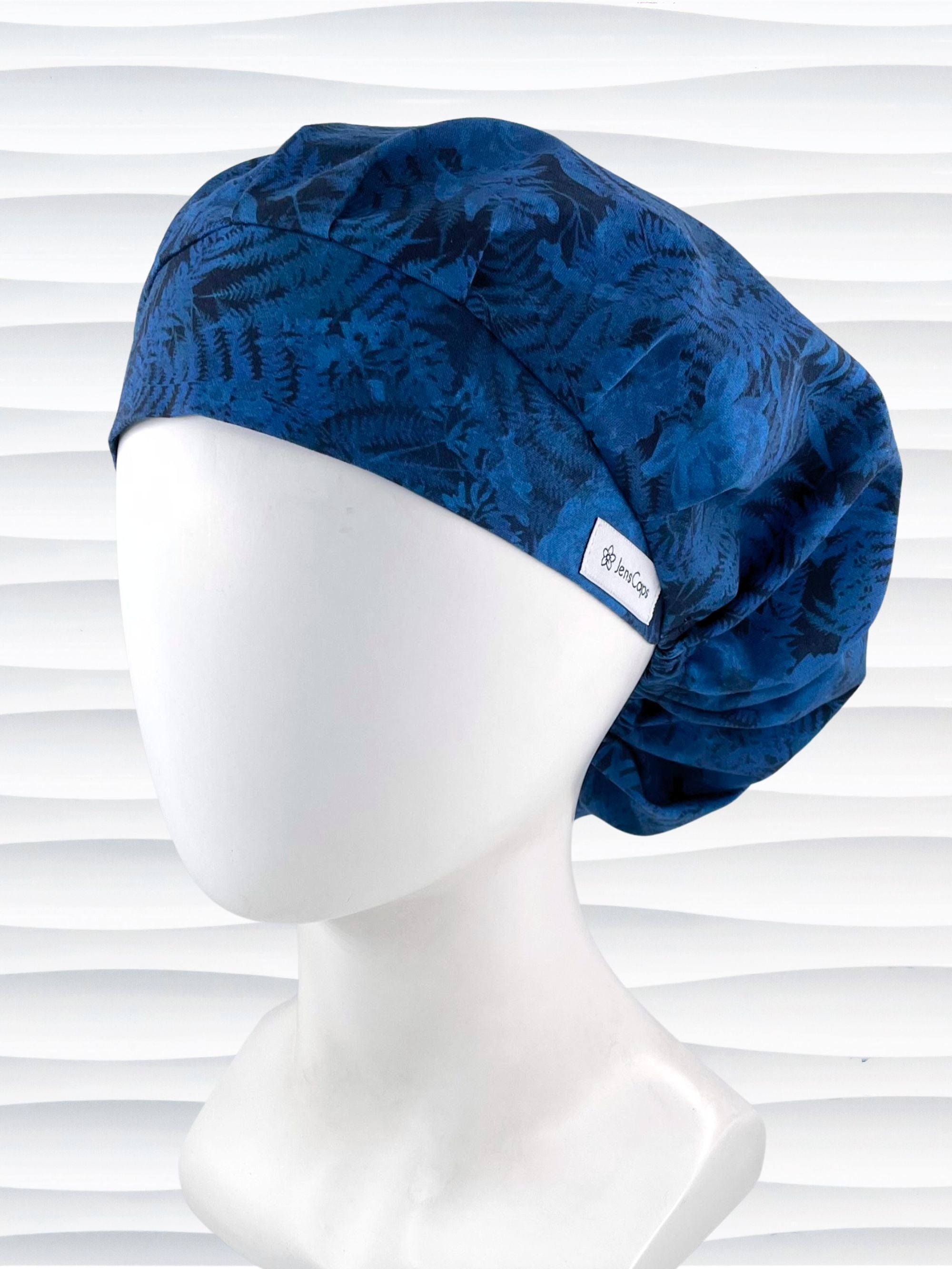 Bouffant style surgical scrub cap with a muted pattern of flowers, ferns, and leaves in dark blue on a deep royal blue cotton fabric.