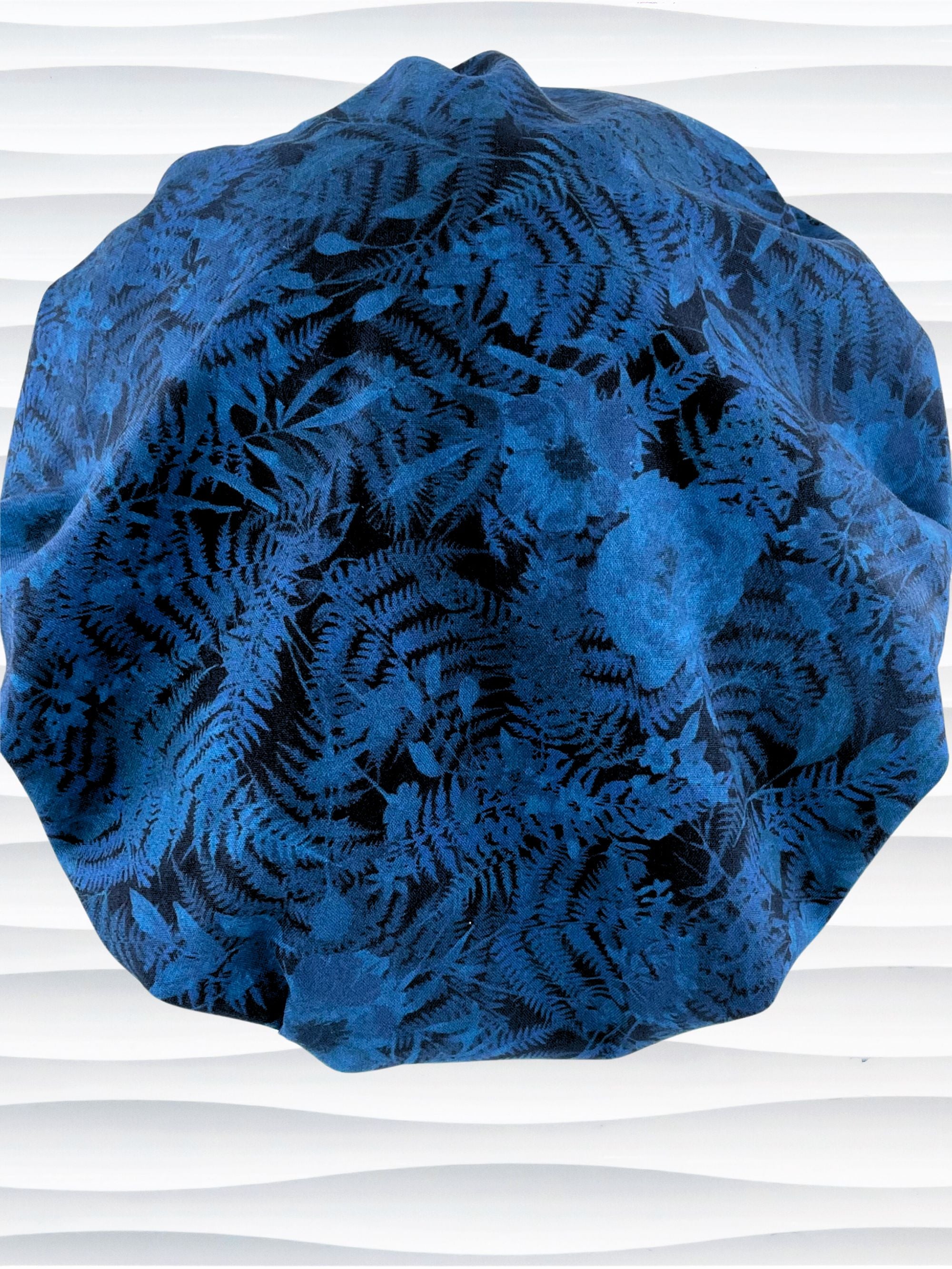 Bouffant style surgical scrub cap with a muted pattern of flowers, ferns, and leaves in dark blue on a deep royal blue cotton fabric.