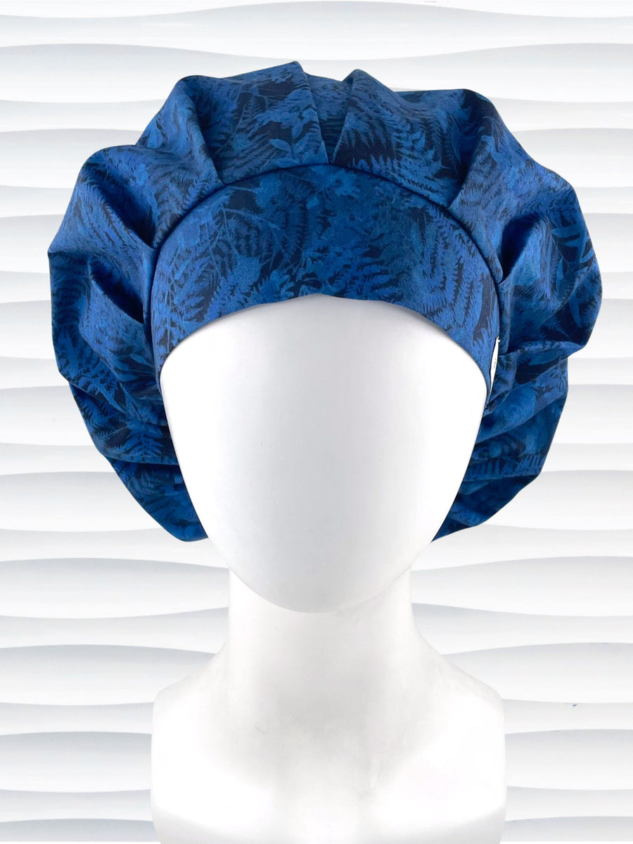 Bouffant style surgical scrub cap with a muted pattern of flowers, ferns, and leaves in dark blue on a deep royal blue cotton fabric.