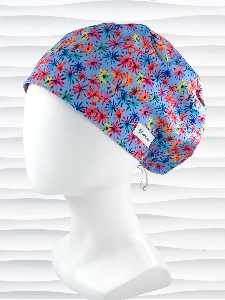 Euro Pixie style surgical scrub cap with bright flowers that look like fireworks in red, pink, yellow, blue, and green all over blue cotton fabric.