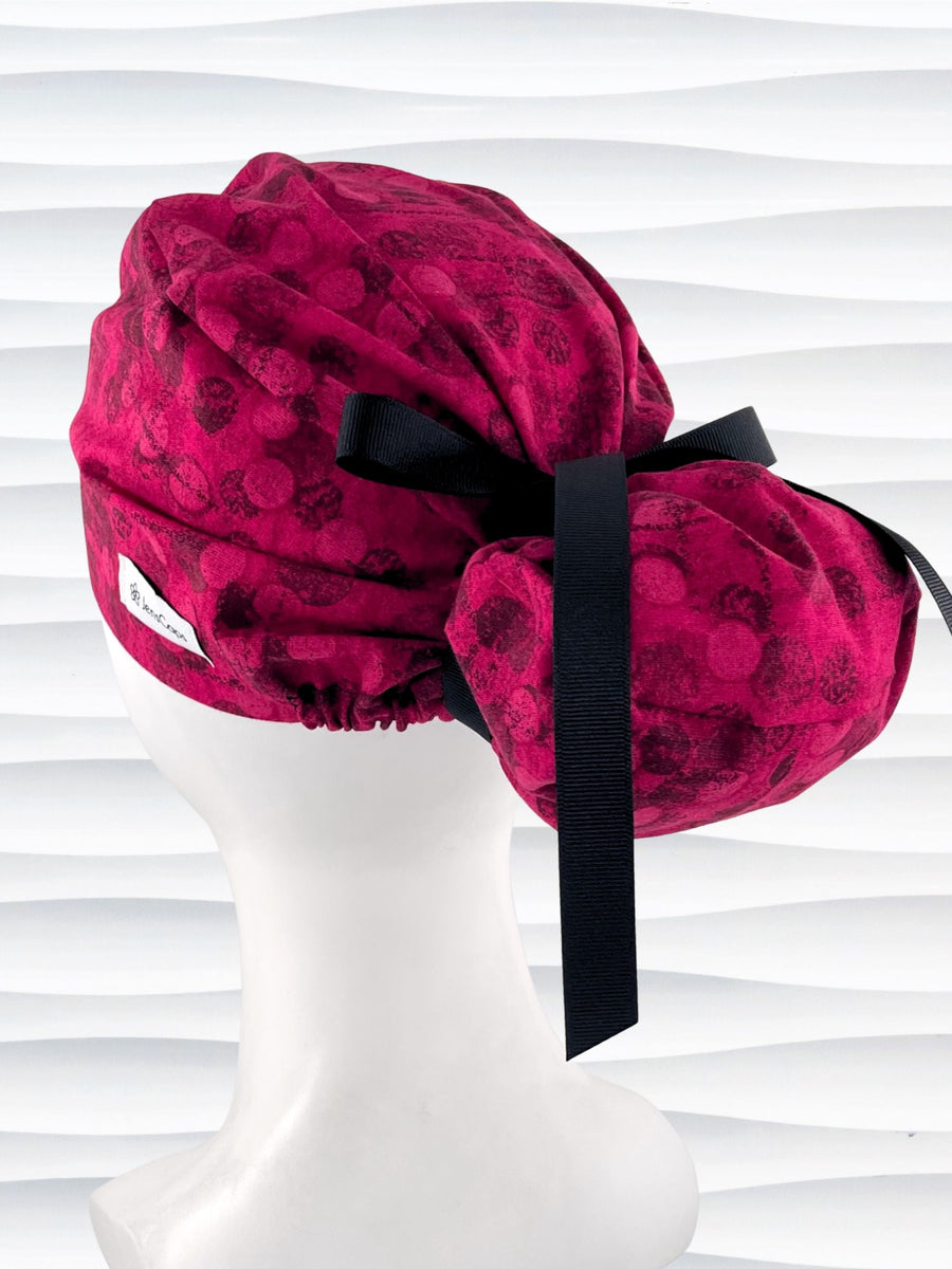 Euro style ponytail surgical scrub cap with a muted pattern of black circles and lines on deep red or magenta cotton fabric.