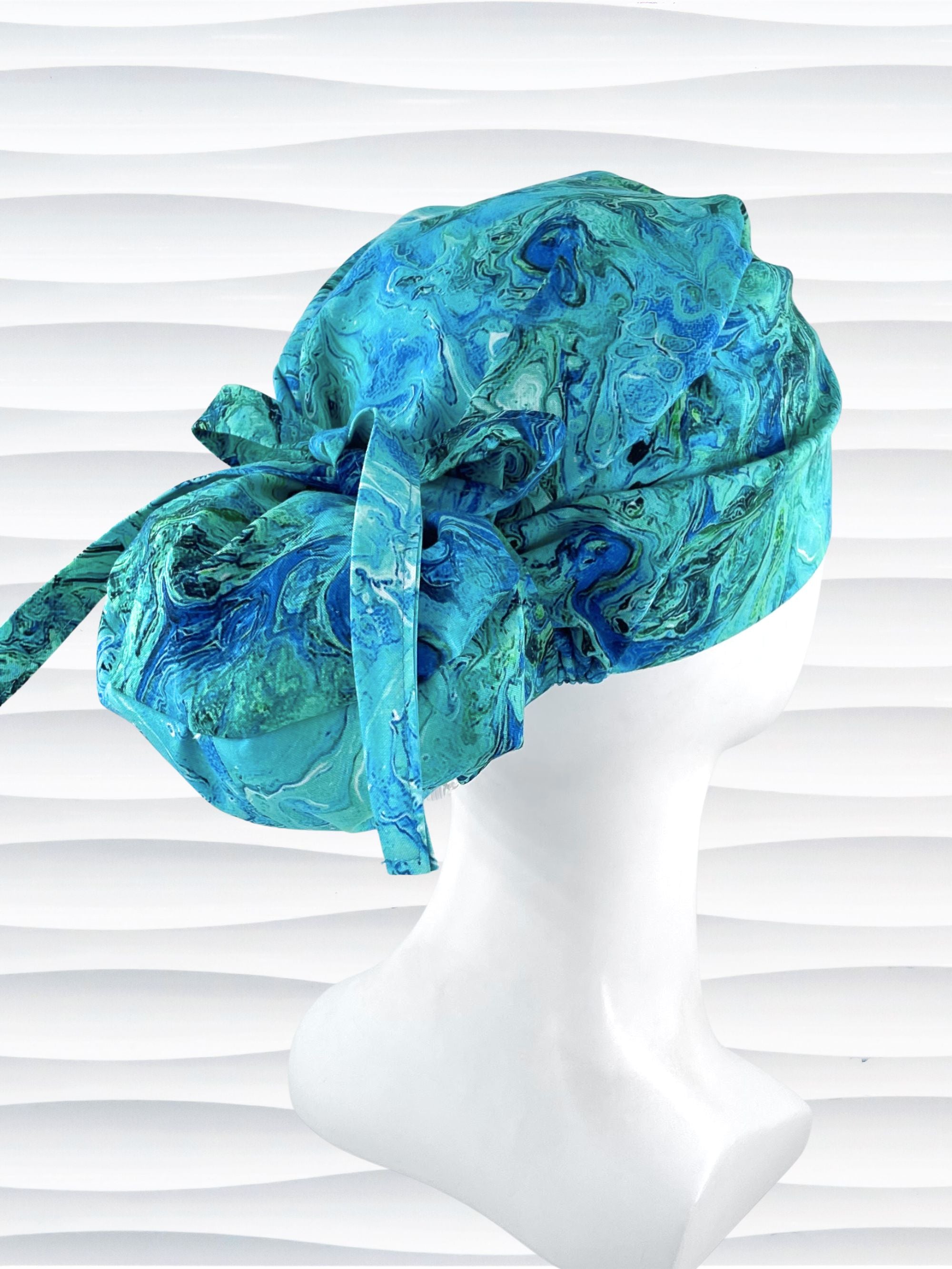 Euro Ponytail surgical scrub cap with a marbled pattern of blues and green that look like ocean tide pools or marbled turquoise on cotton fabric, finished with a tie, and displayed on a white mannequin head.