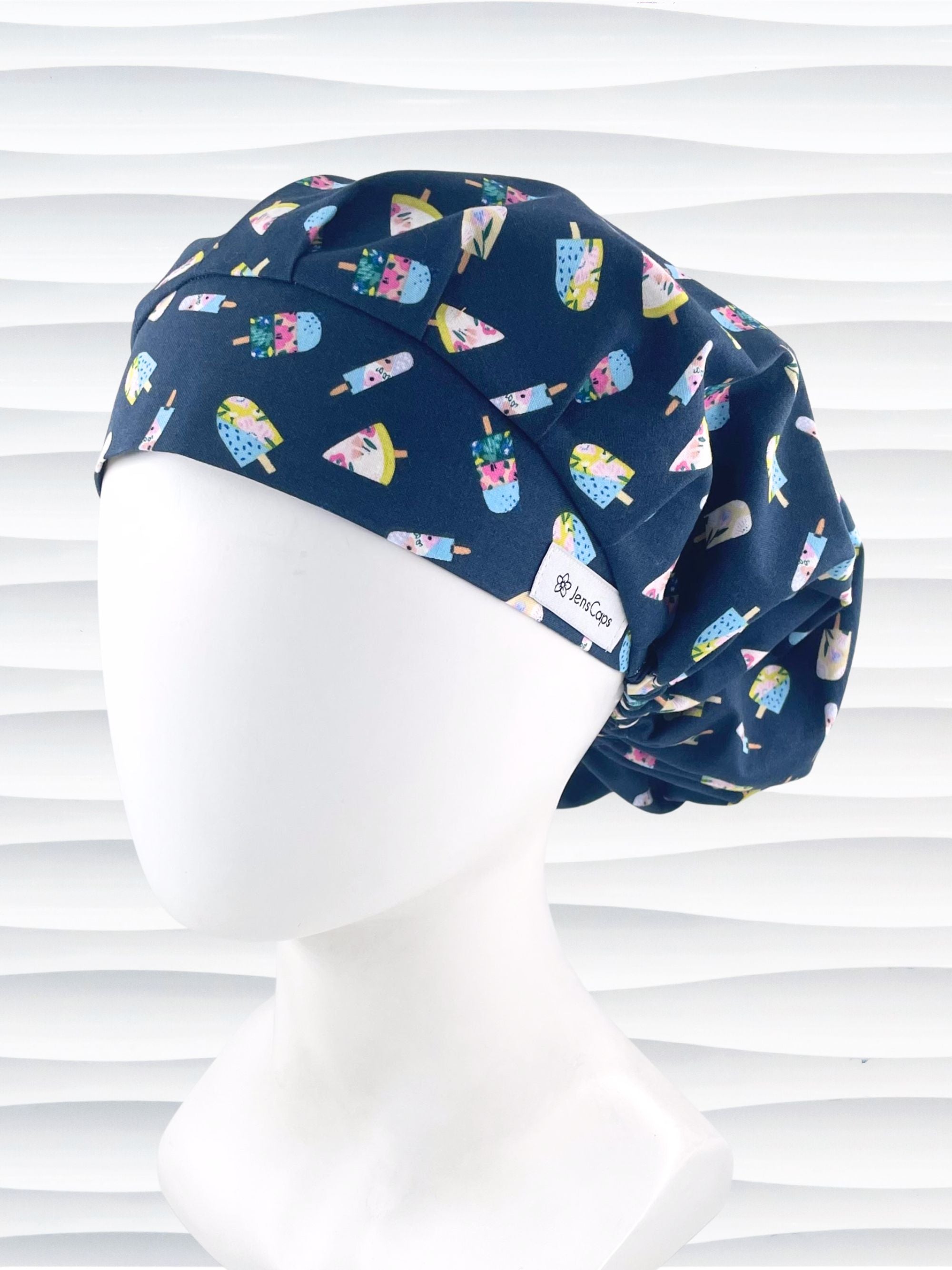 Bouffant style surgical scrub cap side view with popsicles in pink and blue and yellow all over a dark blue cotton fabric.