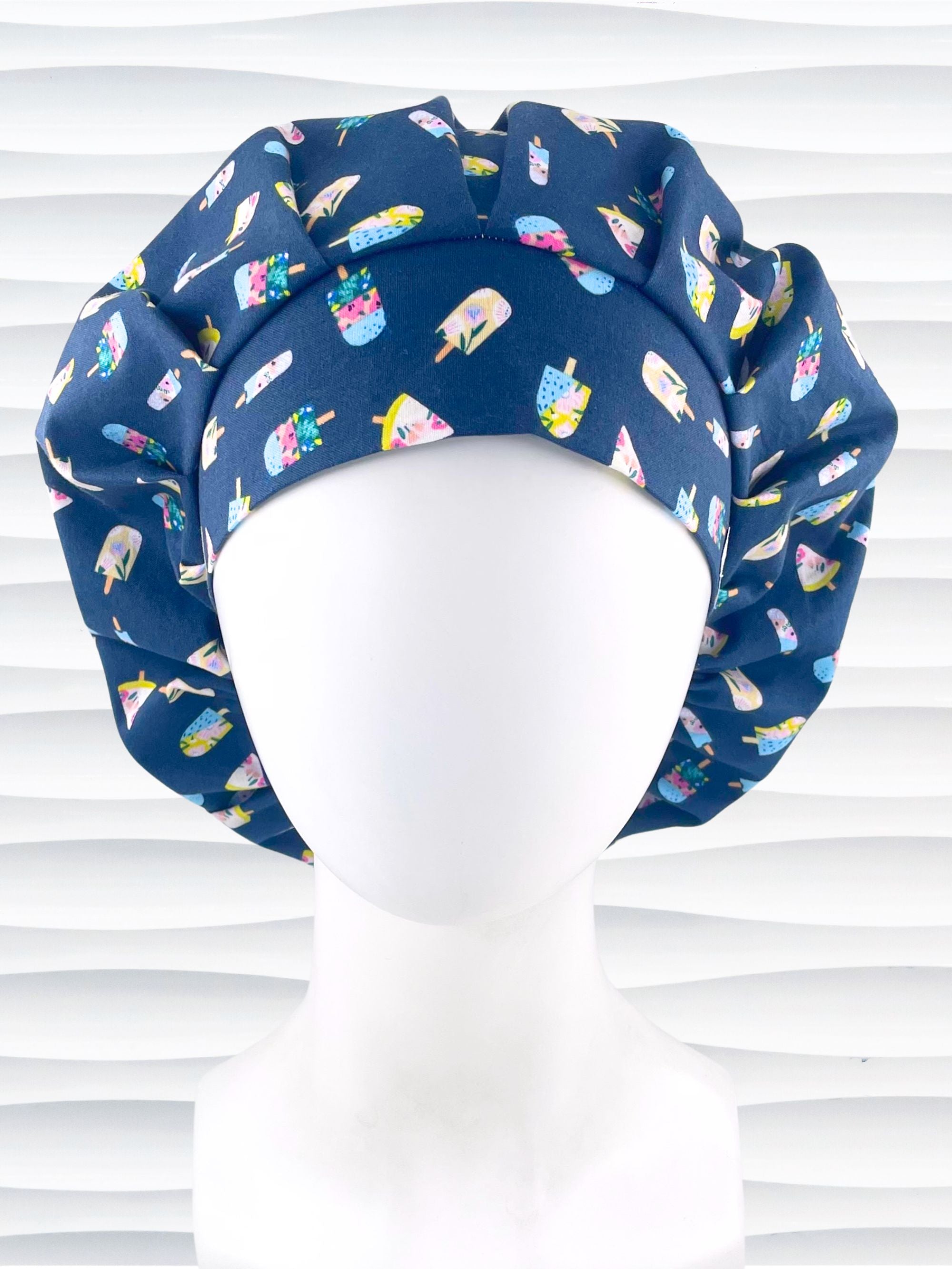 Bouffant style surgical scrub cap front view with popsicles in pink and blue and yellow all over a dark blue cotton fabric.