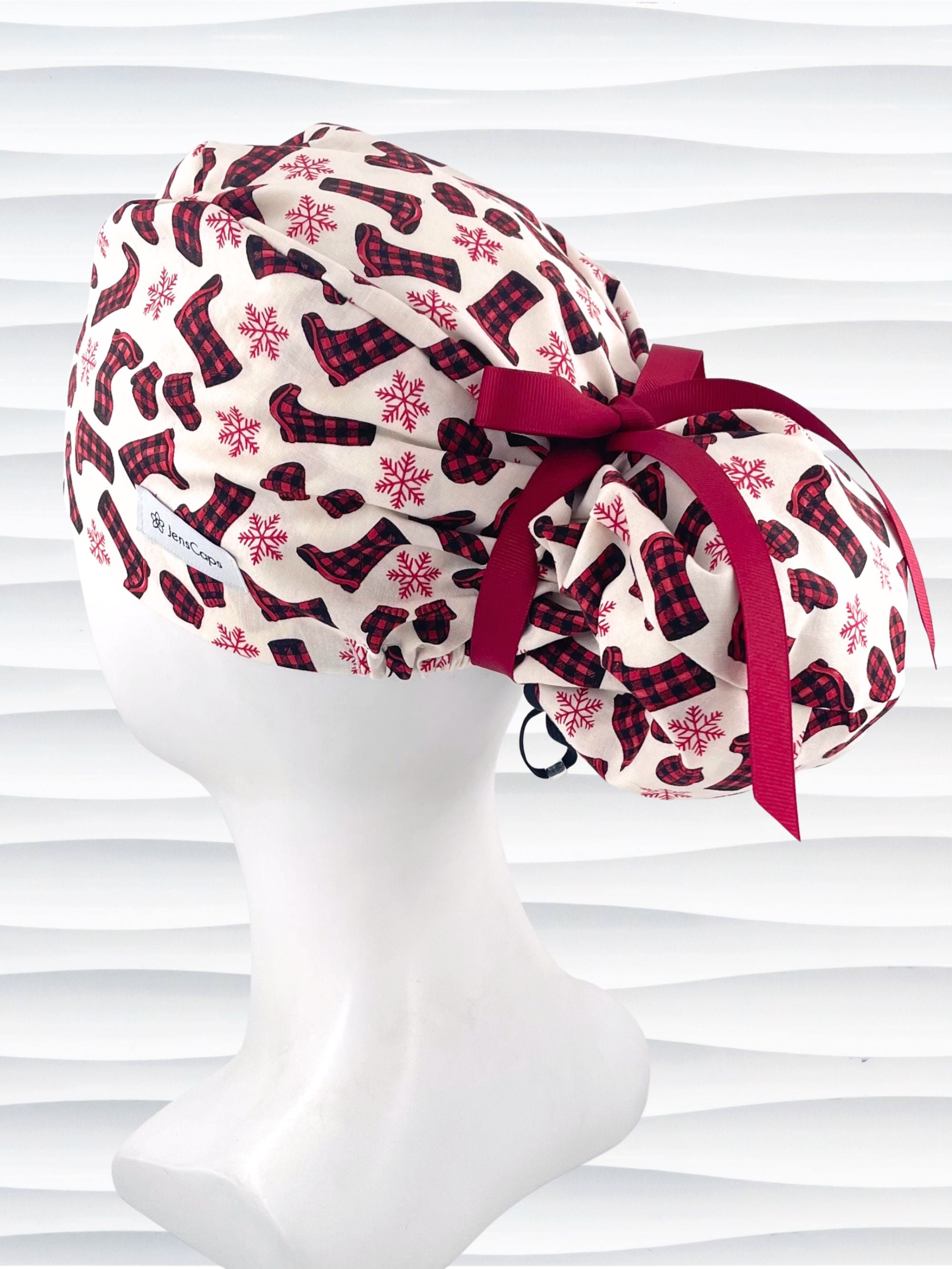 Euro Pony style surgical scrub cap with red snowflakes and red and black buffalo check boots and mittens tossed all over this ivory background on cotton fabric  and finished with a grosgrain ribbon tie.