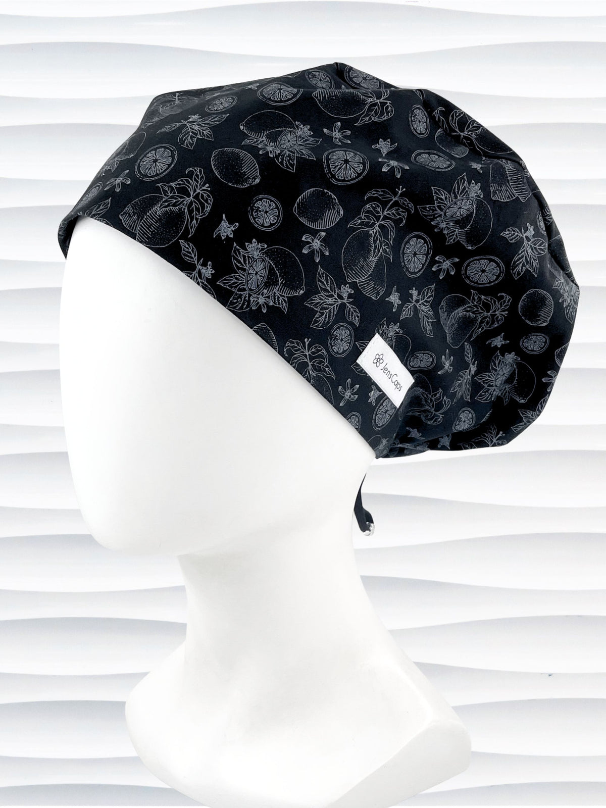Euro Pixie style surgical scrub cap by JensCaps with silver outlines of lemons, lemon leaves, and lemon slices all over a black background and displayed on a white mannequin head.