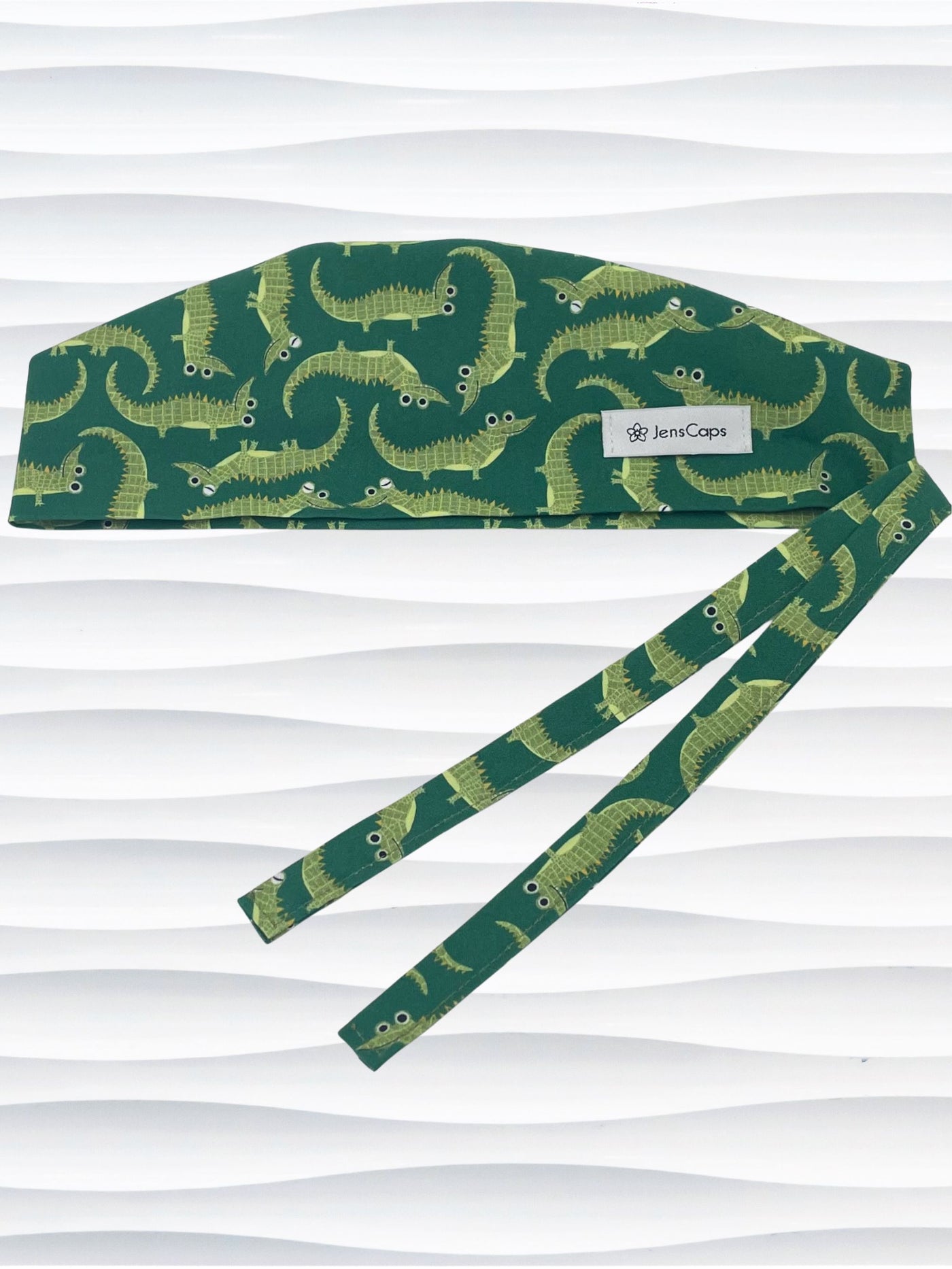 Green surgical scrub cap with playful cartoon alligator print, tied at the back, displayed on a white mannequin head against a textured white background