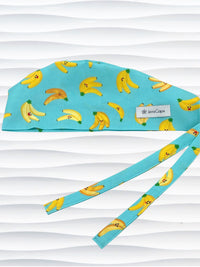Mens style surgical scrub cap with bright yellow bananas tossed on this vibrant blue cotton fabric and finished with adjustable ties shown on mannequin head with a white textured background.
