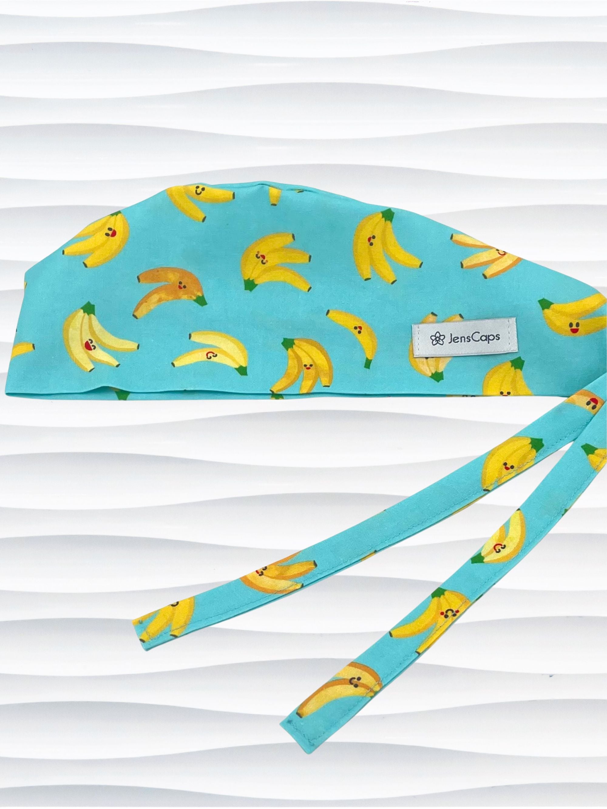 Mens style surgical scrub cap with bright yellow bananas tossed on this vibrant blue cotton fabric and finished with adjustable ties shown on mannequin head with a white textured background.