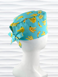 Mens style surgical scrub cap with bright yellow bananas tossed on this vibrant blue cotton fabric and finished with adjustable ties shown on mannequin head with a white textured background.