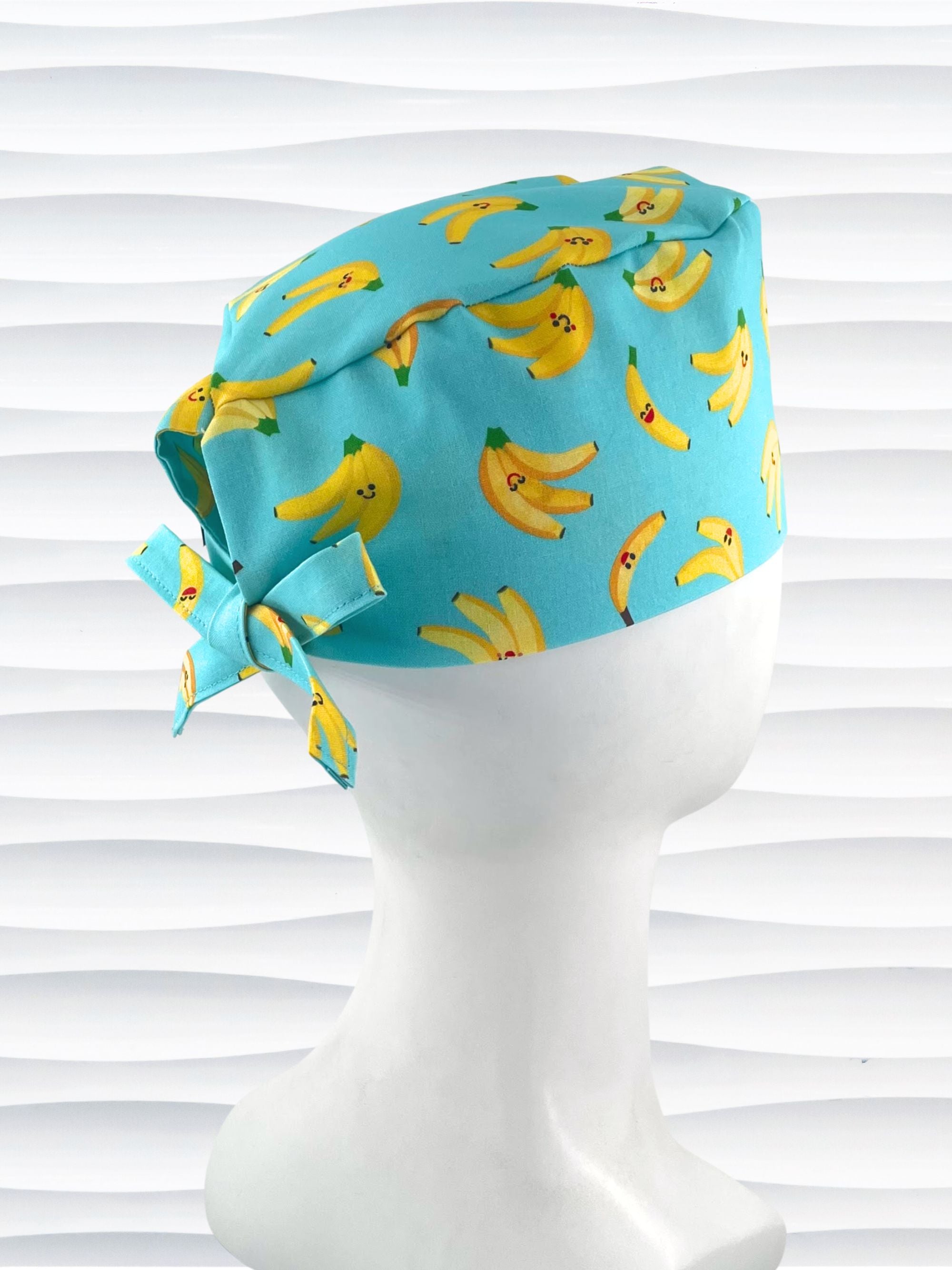 Mens style surgical scrub cap with bright yellow bananas tossed on this vibrant blue cotton fabric and finished with adjustable ties shown on mannequin head with a white textured background.