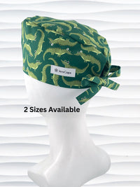 Green surgical scrub cap with playful cartoon alligator print, tied at the back, displayed on a white mannequin head against a textured white background