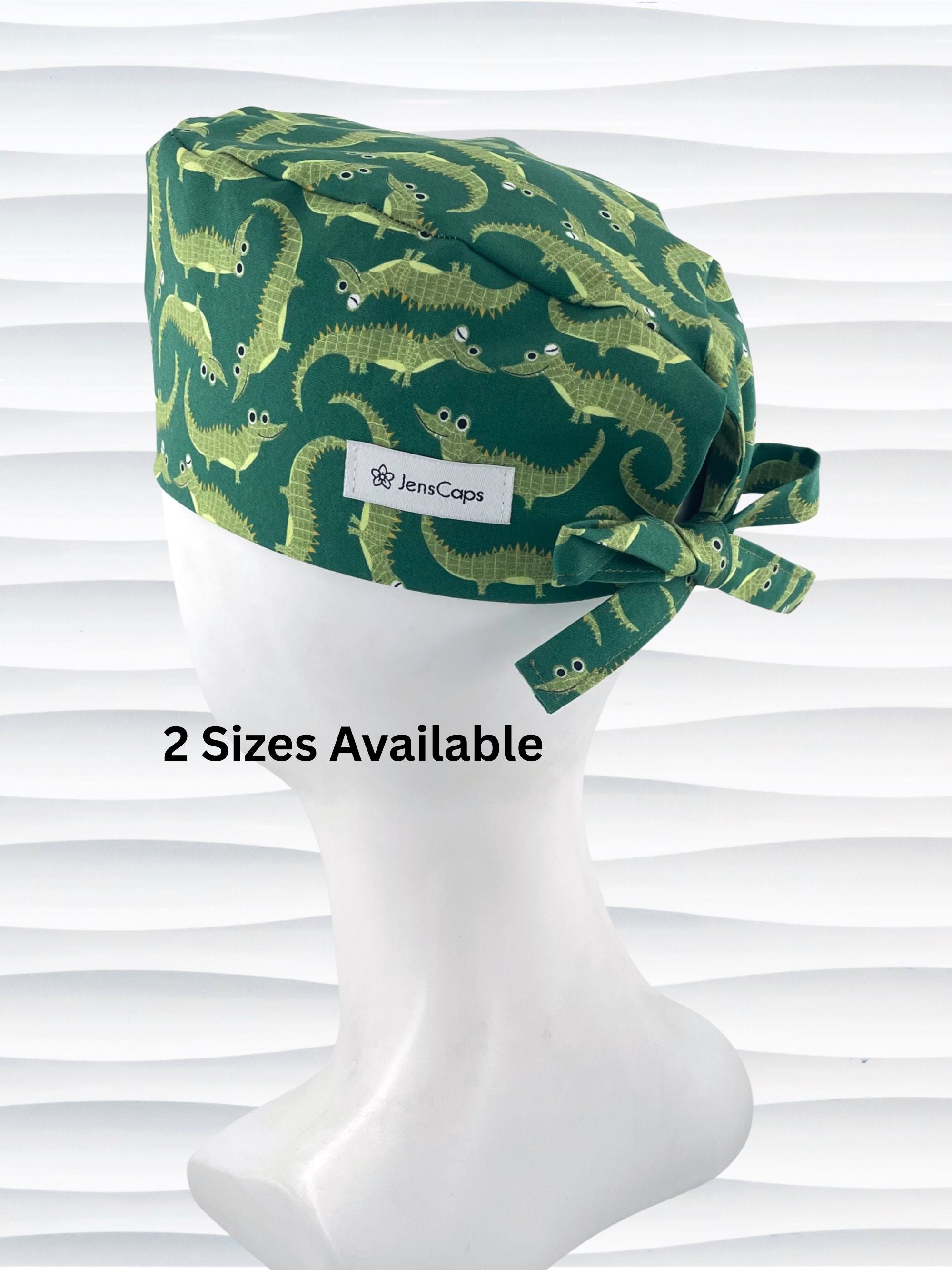 Green surgical scrub cap with playful cartoon alligator print, tied at the back, displayed on a white mannequin head against a textured white background