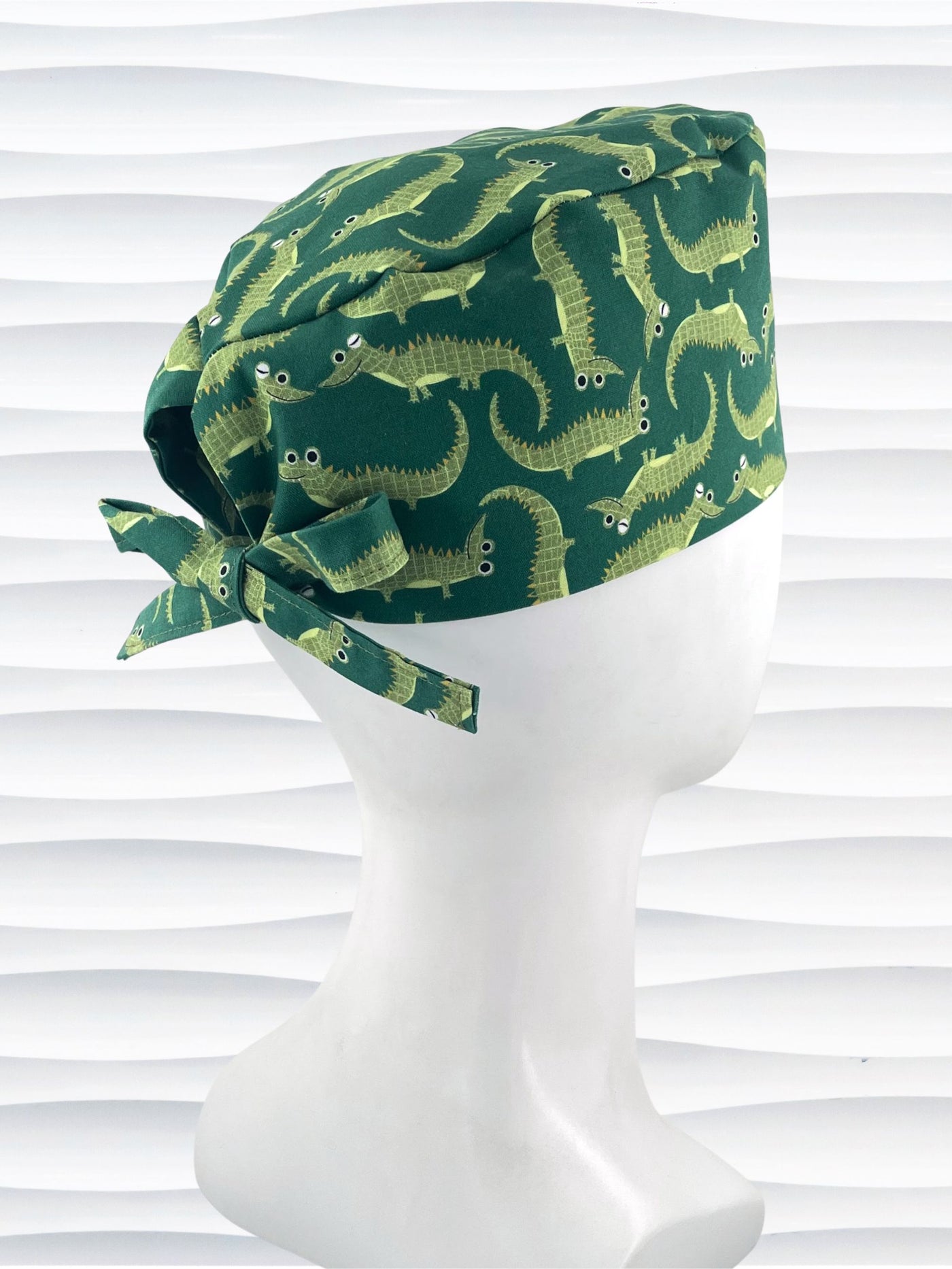 Green surgical scrub cap with playful cartoon alligator print, tied at the back, displayed on a white mannequin head against a textured white background