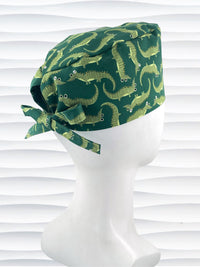 Green surgical scrub cap with playful cartoon alligator print, tied at the back, displayed on a white mannequin head against a textured white background