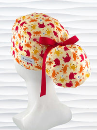 Colorful crab print surgical scrub cap with red ribbon tie, featuring orange, red, and yellow cartoon crabs on cream fabric, displayed on mannequin head – handmade nurse and doctor scrub hat by JensCaps