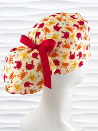 Colorful crab print surgical scrub cap with red ribbon tie, featuring orange, red, and yellow cartoon crabs on cream fabric, displayed on mannequin head – handmade nurse and doctor scrub hat by JensCaps